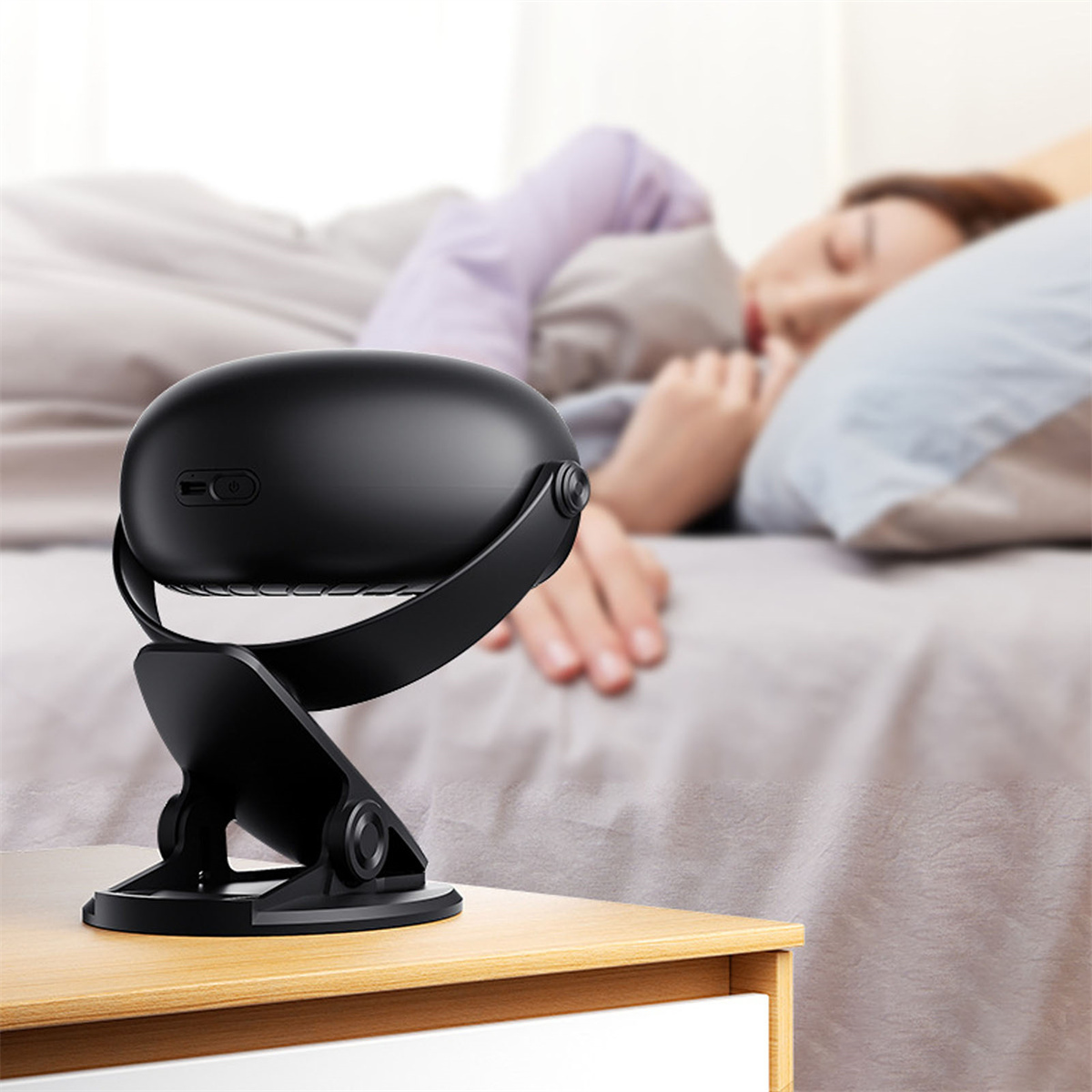 Ein schwarzer Ventilator auf einem Nachttisch neben einer schlafenden Person im Bett.