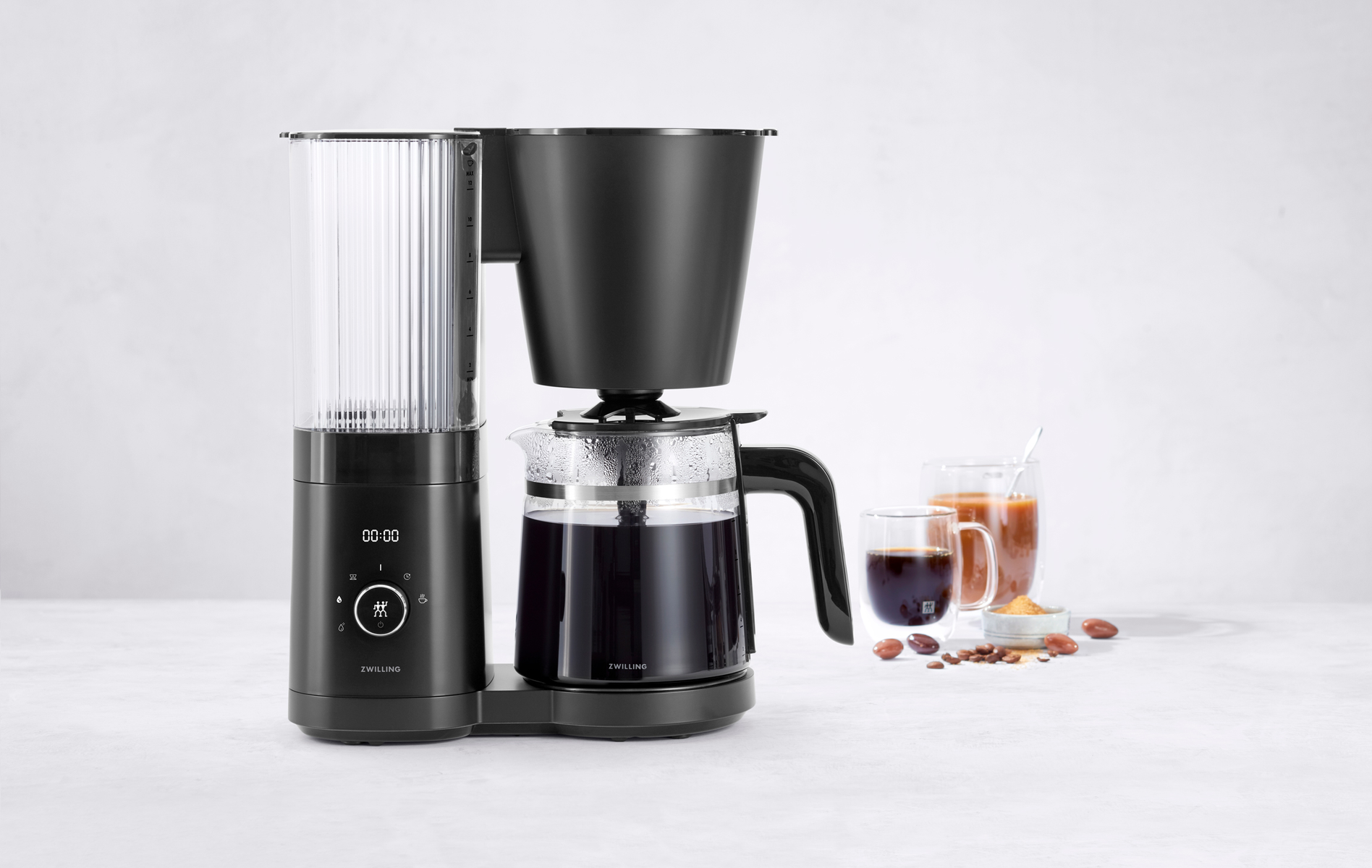 Caffettiera nera e argentata con caraffa in vetro, due bicchieri di caffè e chicchi.