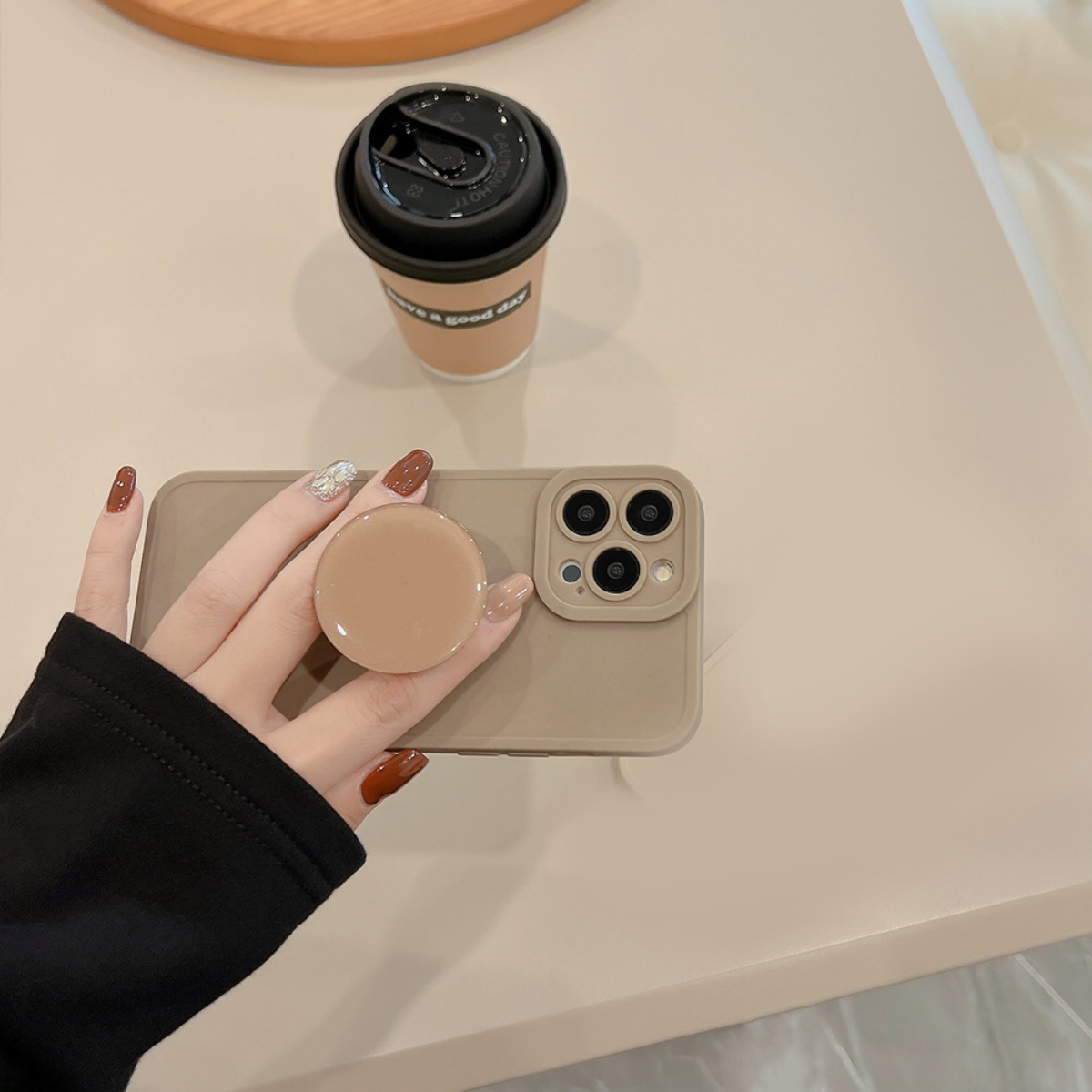 Eine Hand mit manikürten Nägeln, die ein Telefon mit einem beigen PopSocket und einem Kaffeebecher hält.