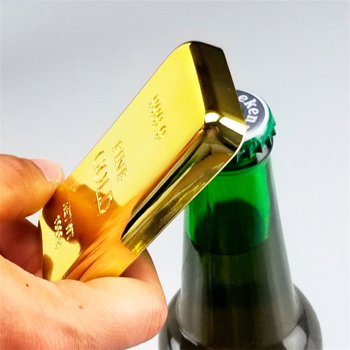 Ein Goldbarren-Flaschenöffner öffnet eine grüne Bierflasche, gehalten von einer Hand.