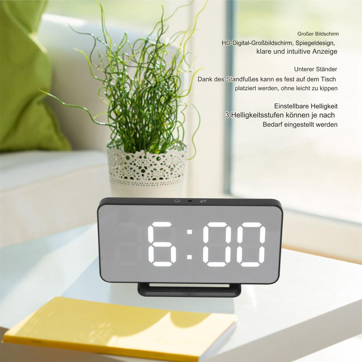 Digitaler Wecker, der 6:00 anzeigt. Er ist schwarz, mit weißen Ziffern, auf einem weißen Tisch, neben einer Pflanze.