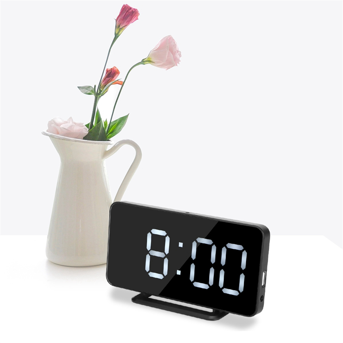 Digitaler Wecker, der 8:00 anzeigt. Er ist schwarz, mit weißen Ziffern, auf einem weißen Tisch, in der Nähe von Blumen in einer Vase.