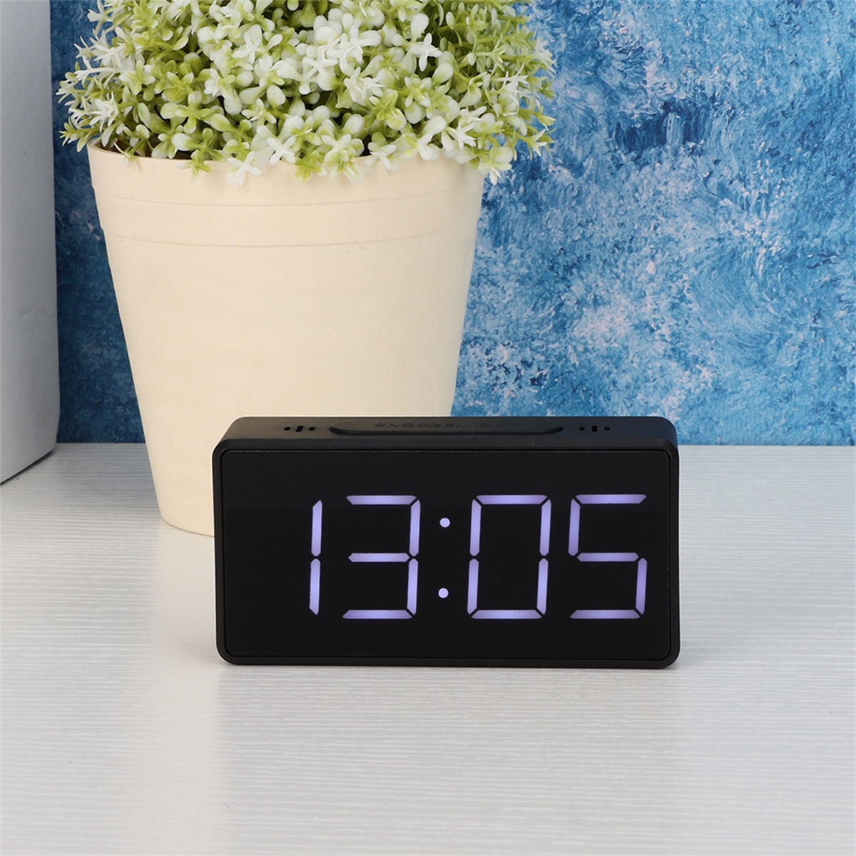 Eine schwarze Digitaluhr, die 13:05 anzeigt, auf einer weißen Oberfläche mit einer Pflanze im Hintergrund platziert.