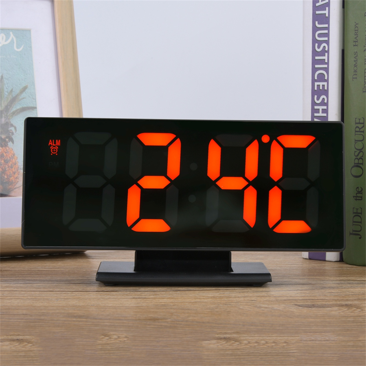 Ein digitaler Wecker zeigt die Uhrzeit 2:40 mit orangefarbenen Ziffern auf schwarzem Hintergrund. Bücher und eine Palme im Hintergrund.