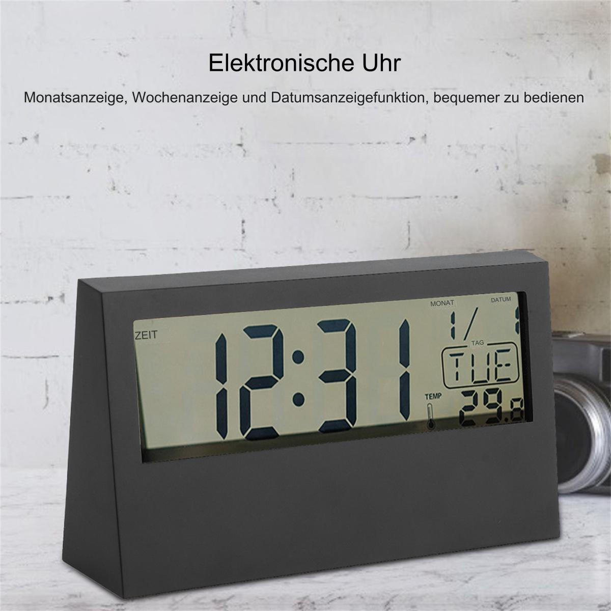 Eine schwarze Digitaluhr, die Zeit, Datum und Wochentag auf einer hellen Oberfläche anzeigt.