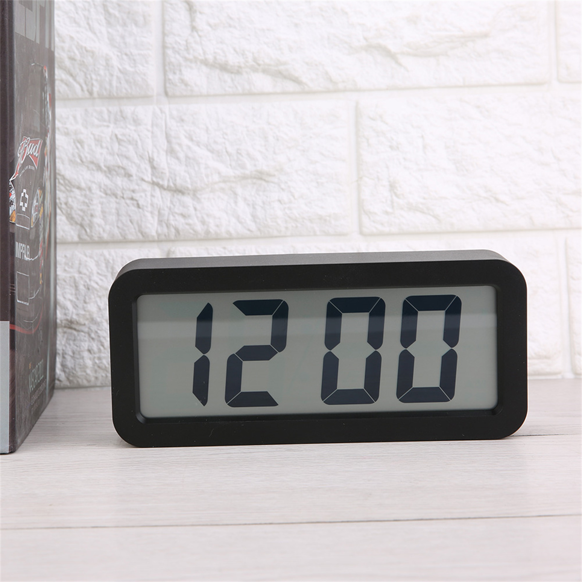 Schwarze Digitaluhr mit der Anzeige '12:00' auf einem weißen Bildschirm, auf einer Holzoberfläche.