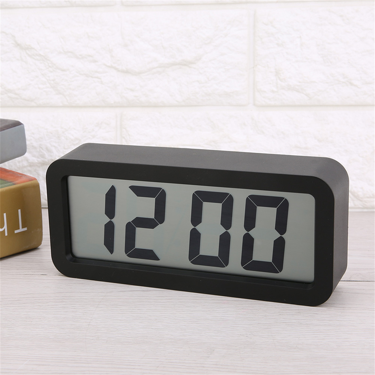 Schwarzer digitaler Wecker mit der Anzeige 12:00. Der Hintergrund ist eine weiße Ziegelmauer.