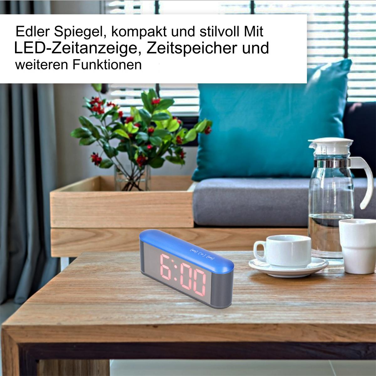Ein blauer digitaler Wecker auf einem Holztisch. Eine Pflanze und eine Tasse stehen daneben.