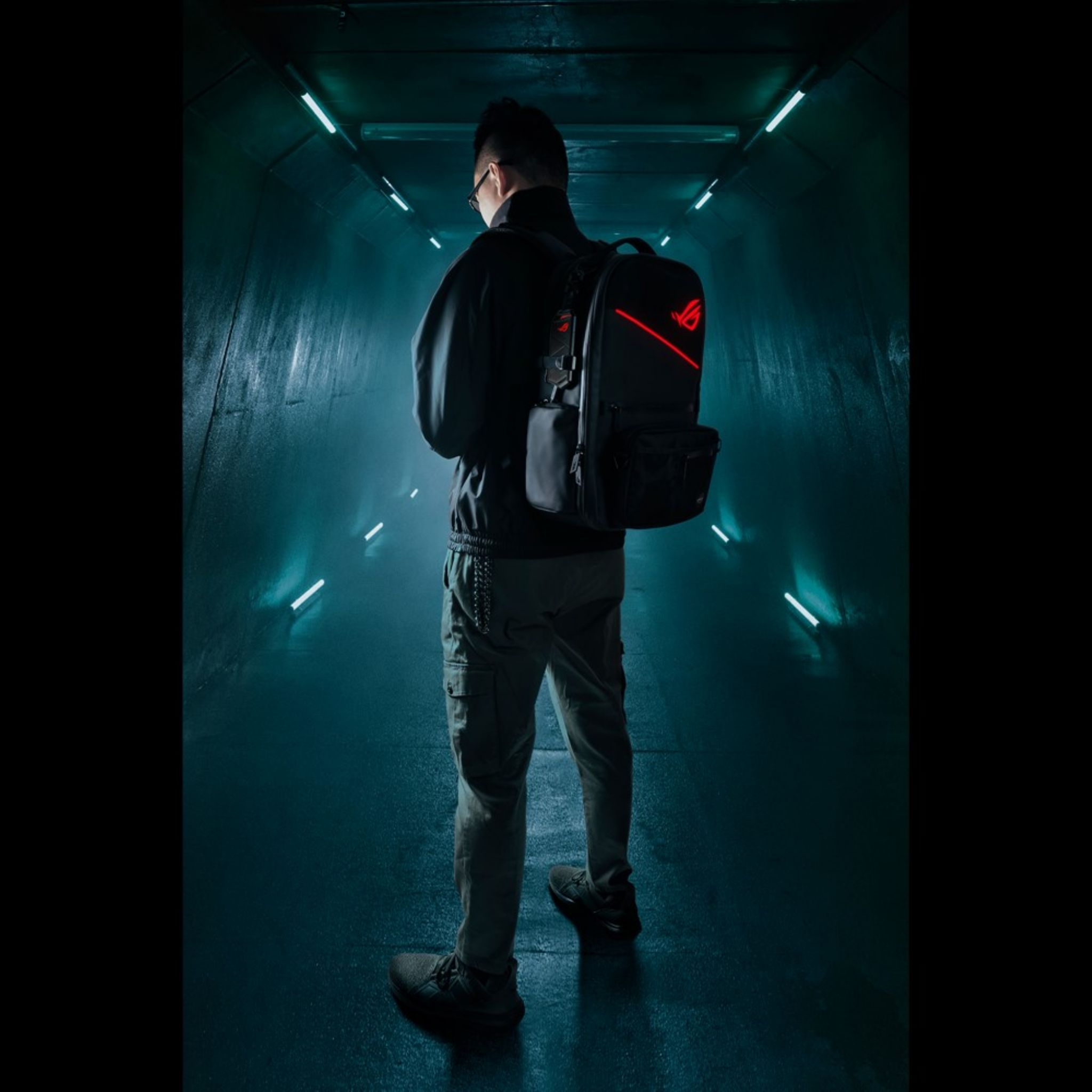 Mężczyzna w ciemnych ubraniach stoi w świecącym tunelu, w okularach i z plecakiem z czerwonym logo.