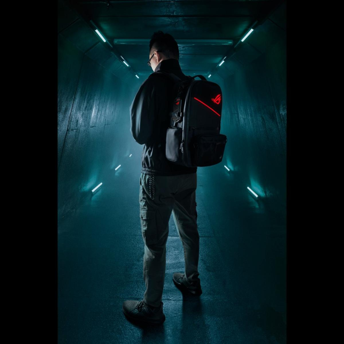 Mężczyzna w ciemnym tunelu z plecakiem. Nosi okulary, czarną kurtkę, spodnie cargo i czarne buty. Plecak ma czerwone logo.