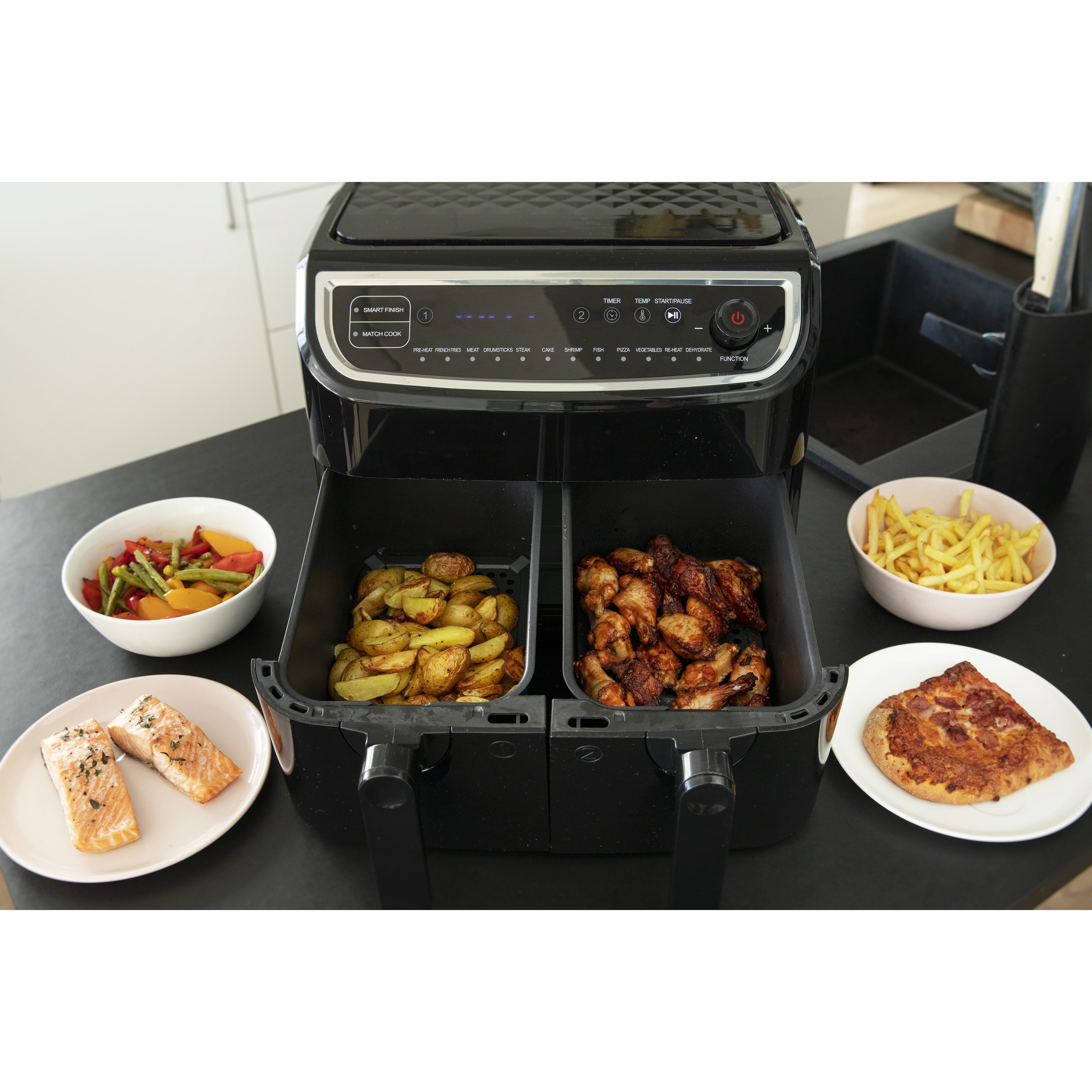 Zwarte airfryer met eten: aardappelen, kippenvleugels, pizza, groenten en zalm op borden.