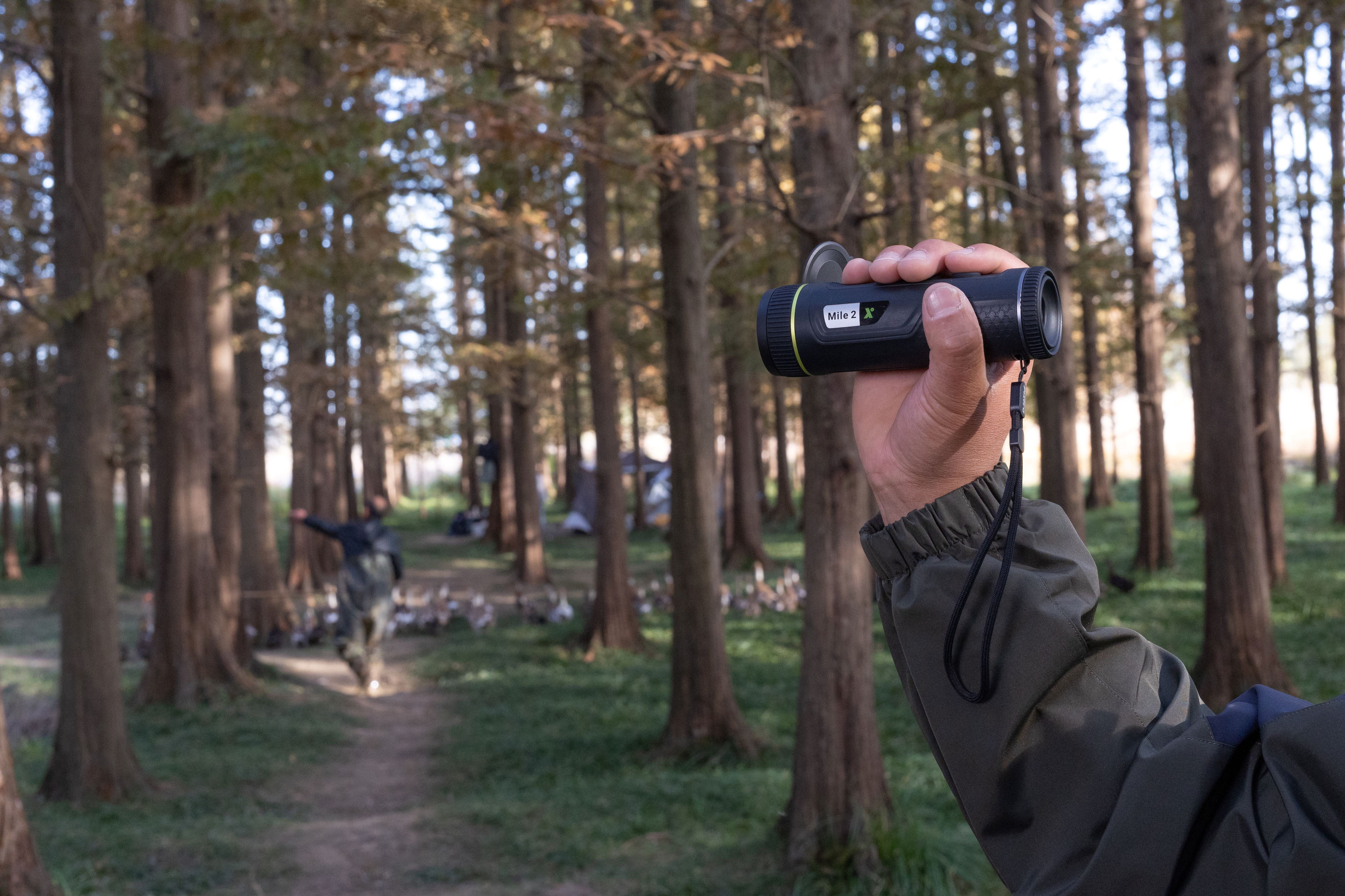 Persona en un bosque, sosteniendo un monocular, con árboles y otra persona detrás.