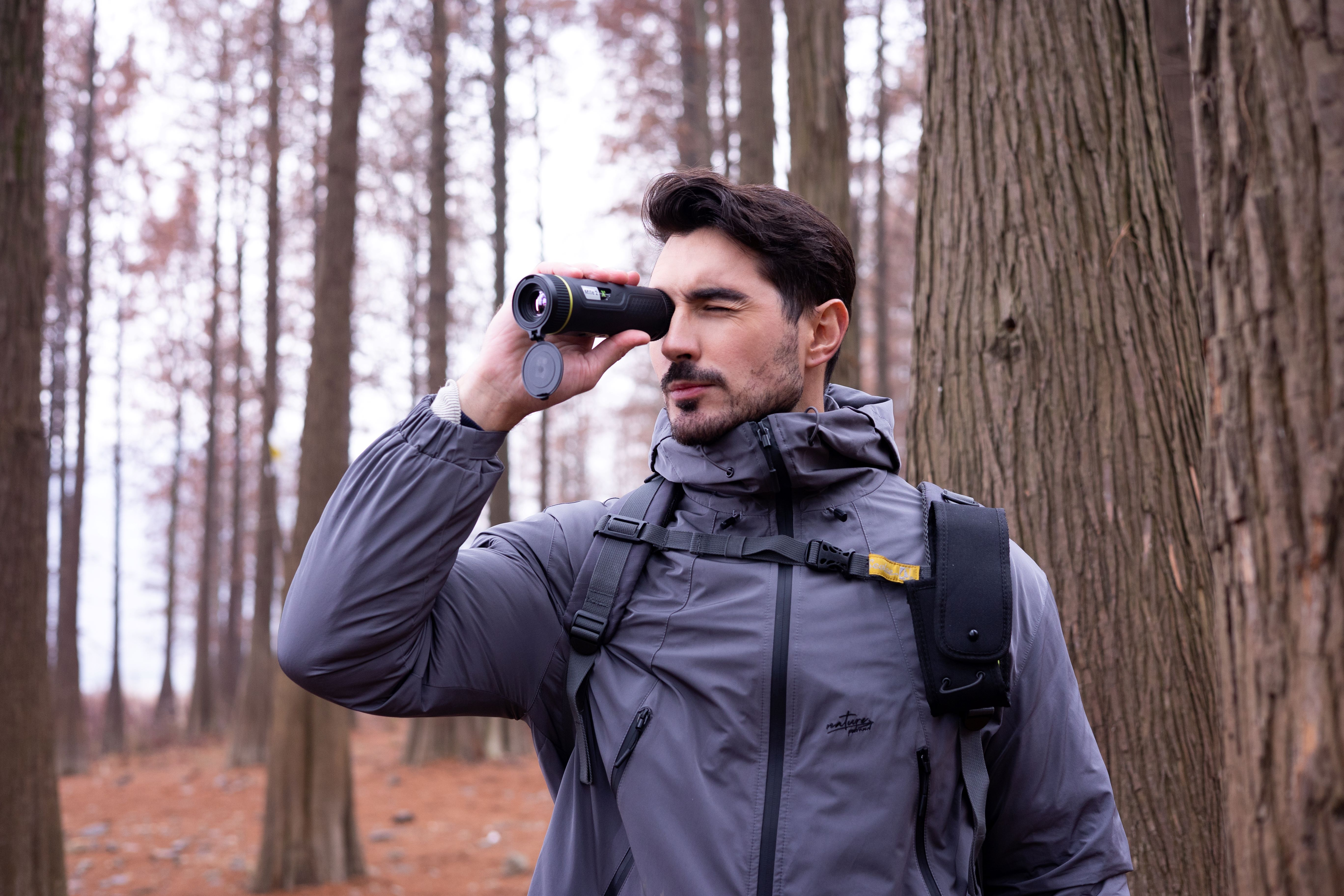 Hombre usando un monocular en un bosque, con una chaqueta gris y una mochila.