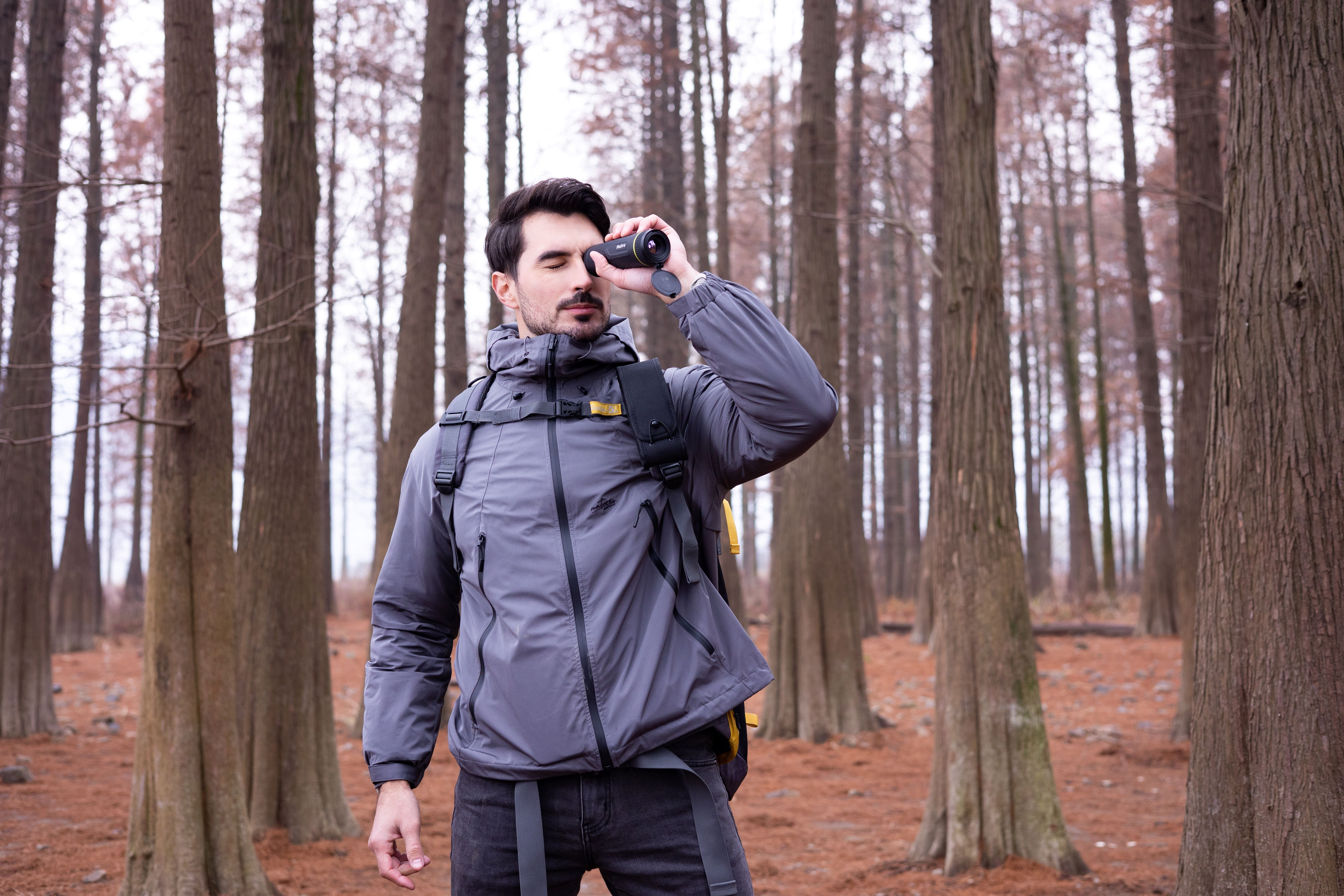 Un hombre en un bosque mirando a través de binoculares. Árboles altos en el fondo. Cielo nublado.