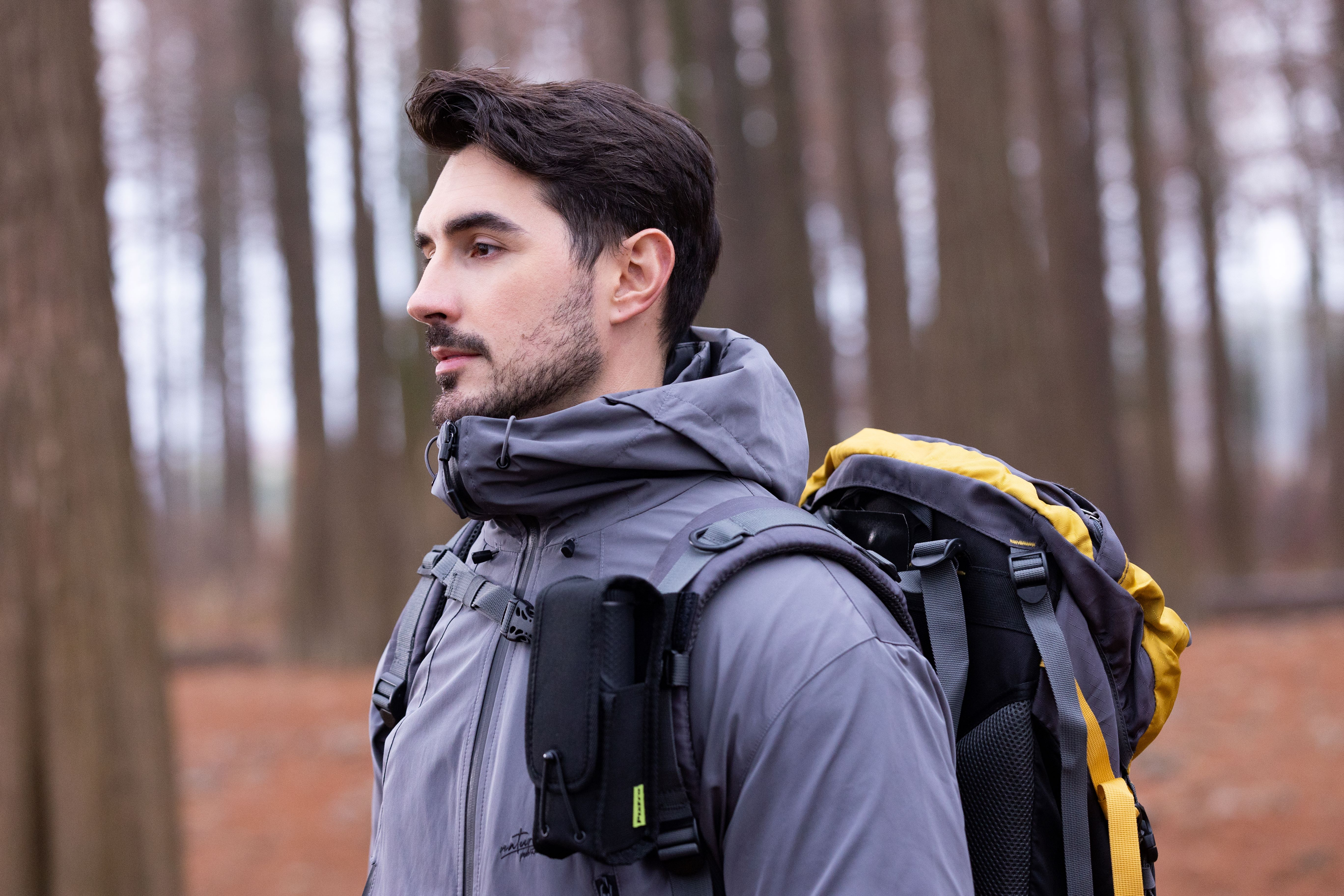 Hombre con mochila camina al aire libre, con chaqueta gris, árboles al fondo.