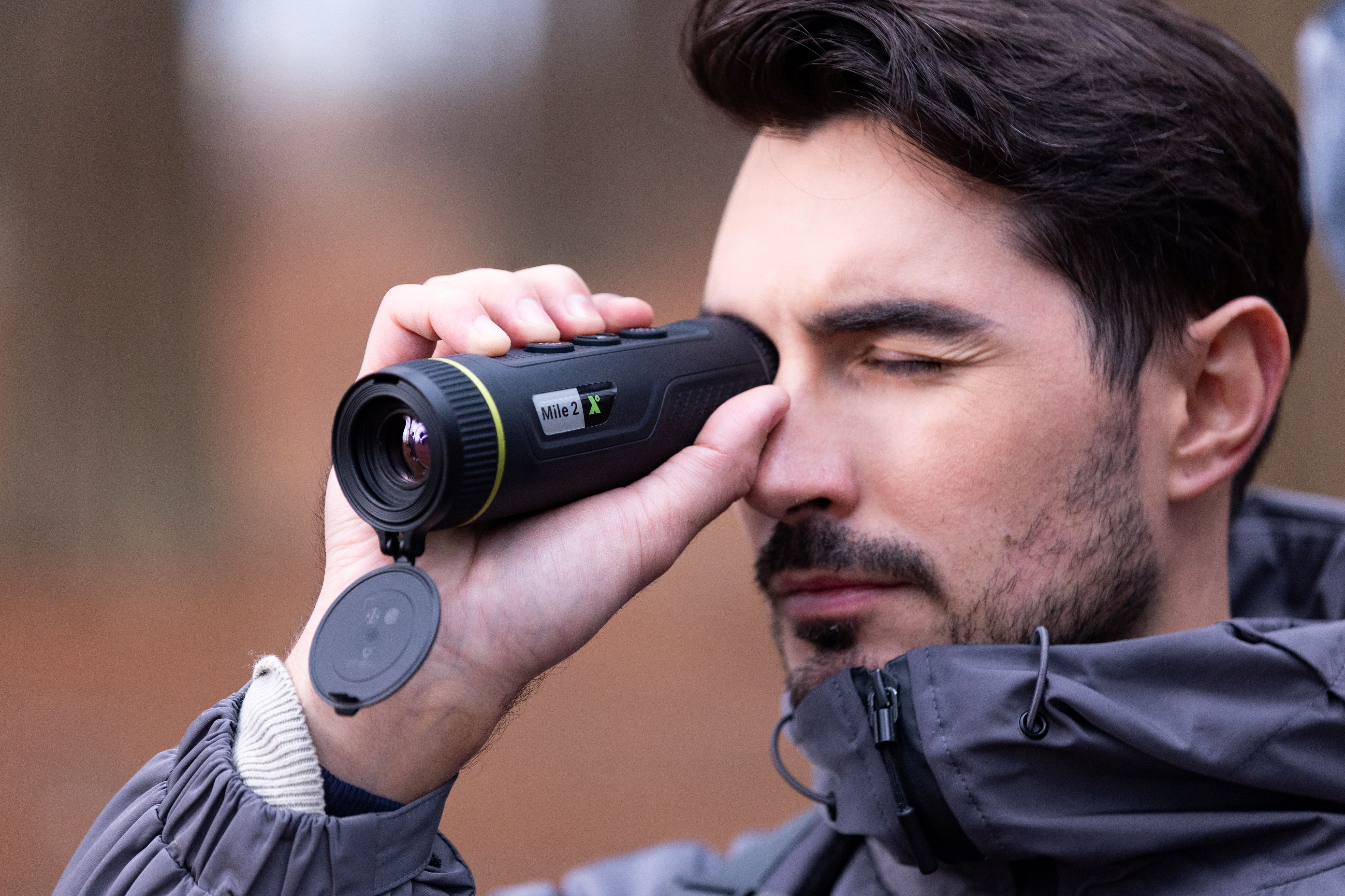 Un hombre mirando a través de un monocular negro. Está afuera. Fondo verde y marrón.