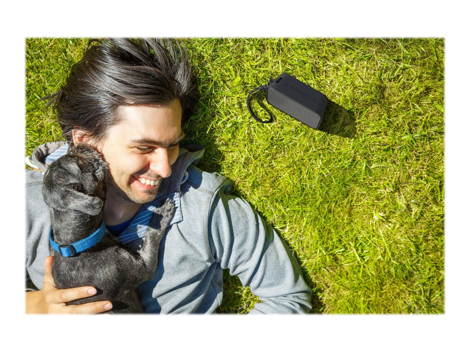 Hombre acostado en la hierba, sonriendo, abrazando a un perro negro pequeño, y un altavoz negro cerca.