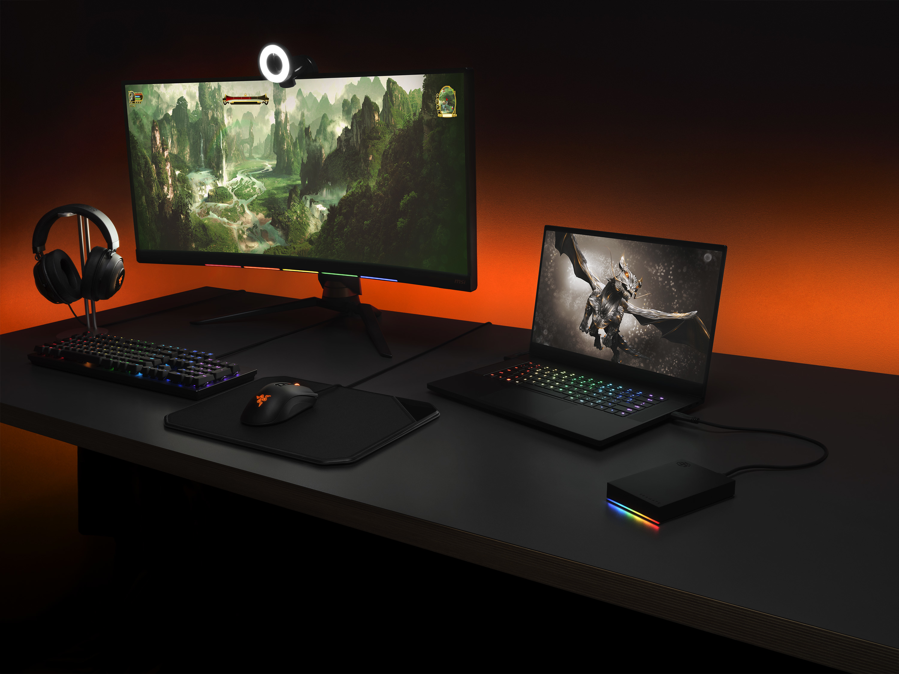 Gaming-Setup mit Laptop, Monitor, Tastatur, Maus und Kopfhörern. Die Szene ist mit orangefarbenem Licht beleuchtet.