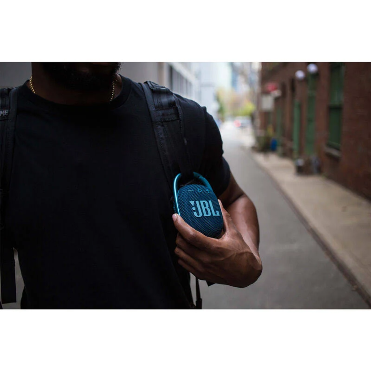 Un hombre con una mochila sosteniendo un pequeño altavoz JBL azul, caminando por un callejón.