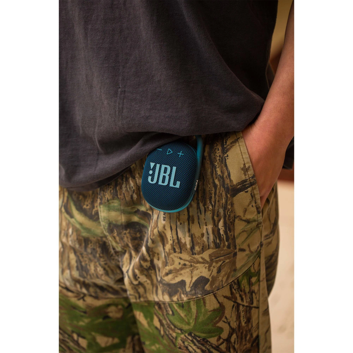 Persona con camisa gris y pantalones camuflaje, sosteniendo un altavoz JBL azul en el bolsillo.