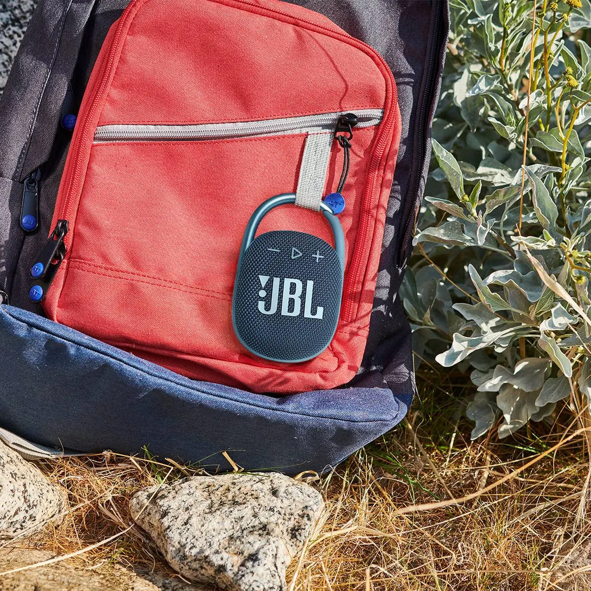 Altavoz JBL Clip sujeto a una mochila, cerca de plantas y rocas.