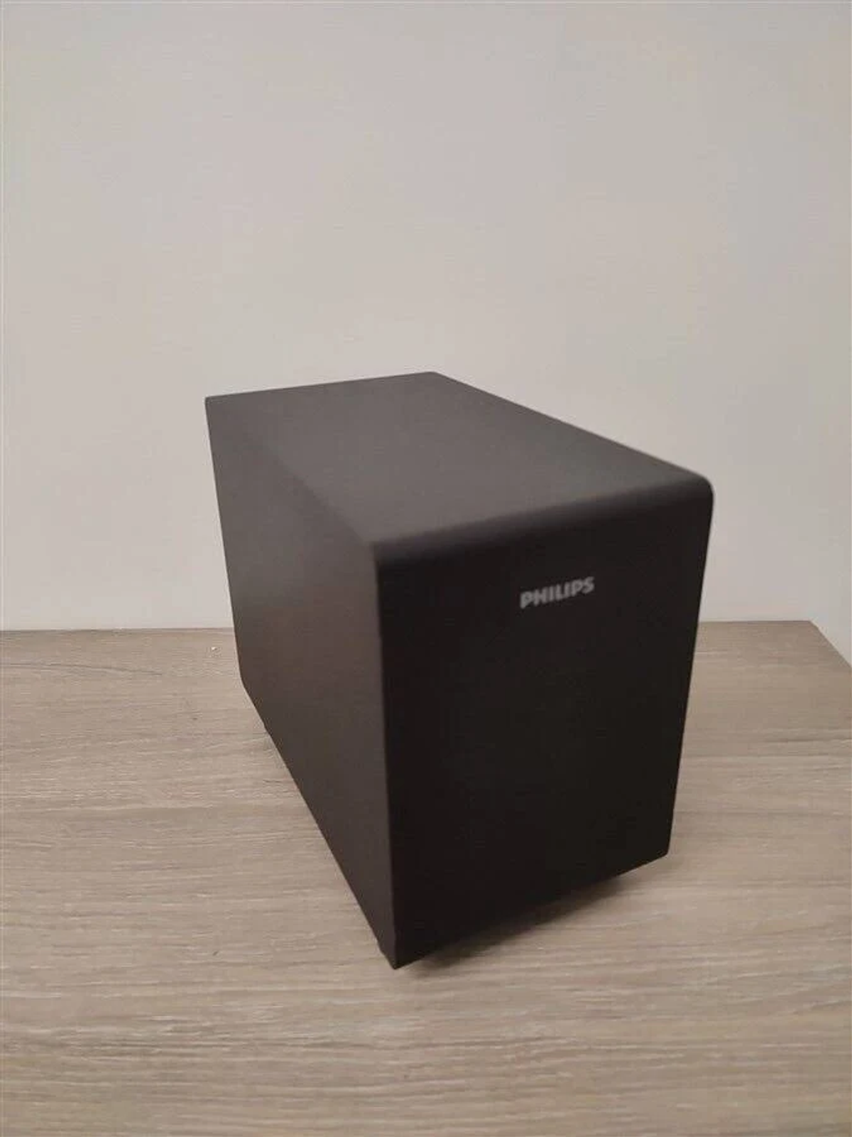 Czarny subwoofer Philips stoi na jasnobrązowej drewnianej powierzchni na tle białej ściany.