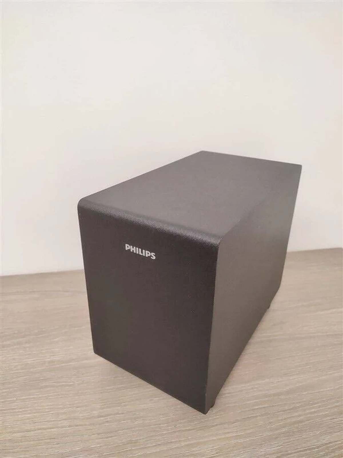 Czarny głośnik subwoofera na drewnianej powierzchni na tle białej ściany.