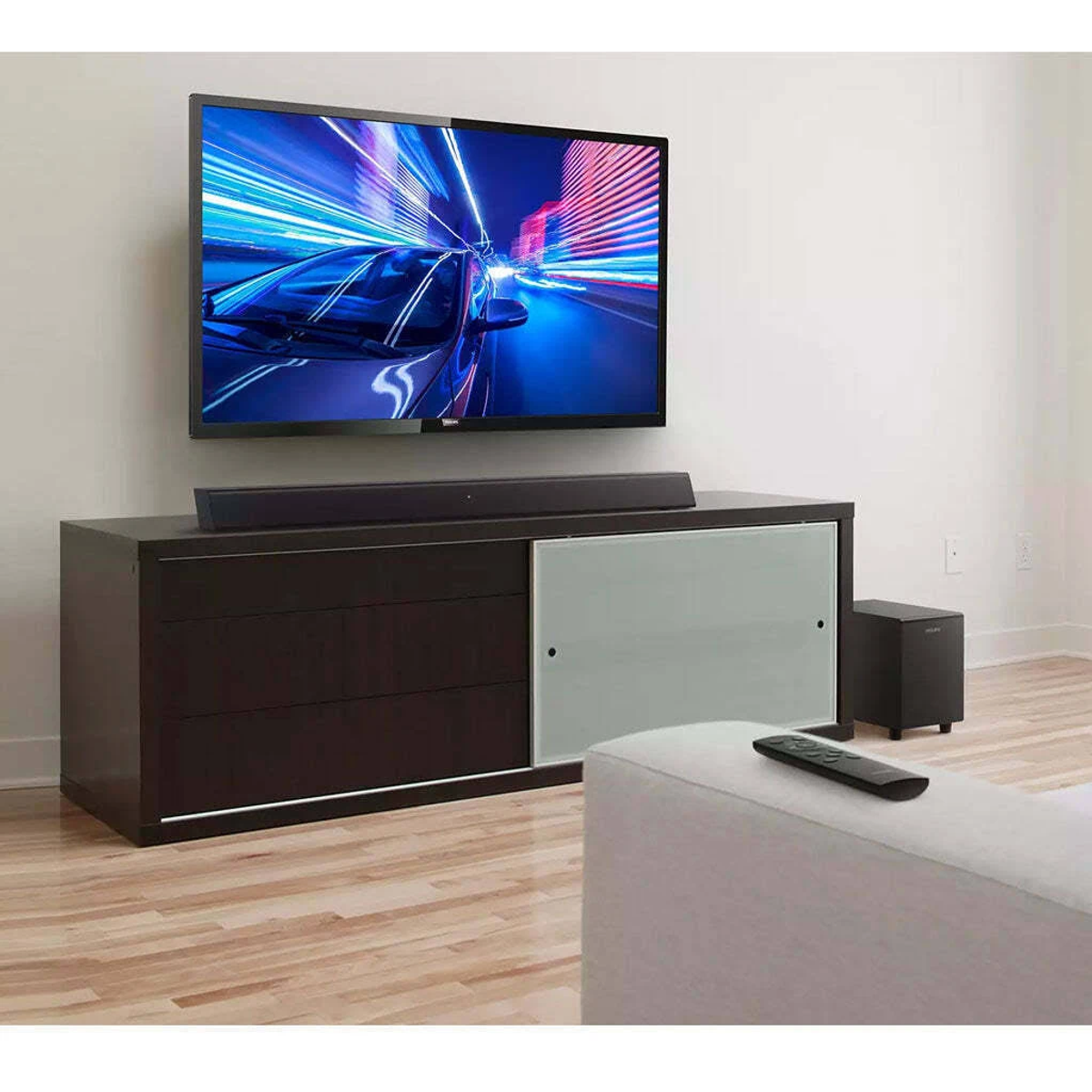 Telewizor na ciemnej szafce z soundbarem i subwooferem. Widoczna biała sofa i pilot.