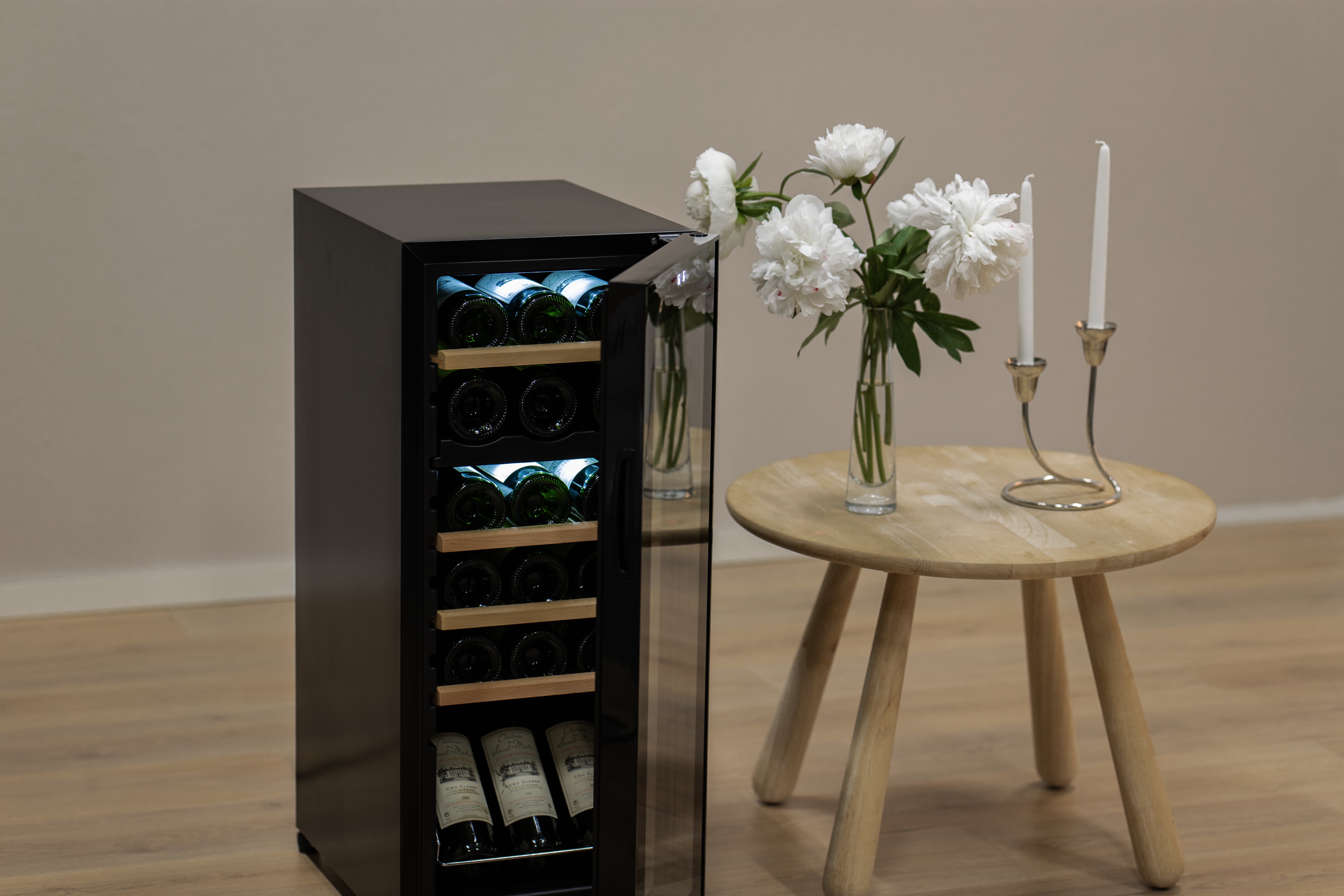 Ein schwarzer Weinkühlschrank mit Flaschen im Inneren und einer Glastür. Daneben stehen Blumen und Kerzen.