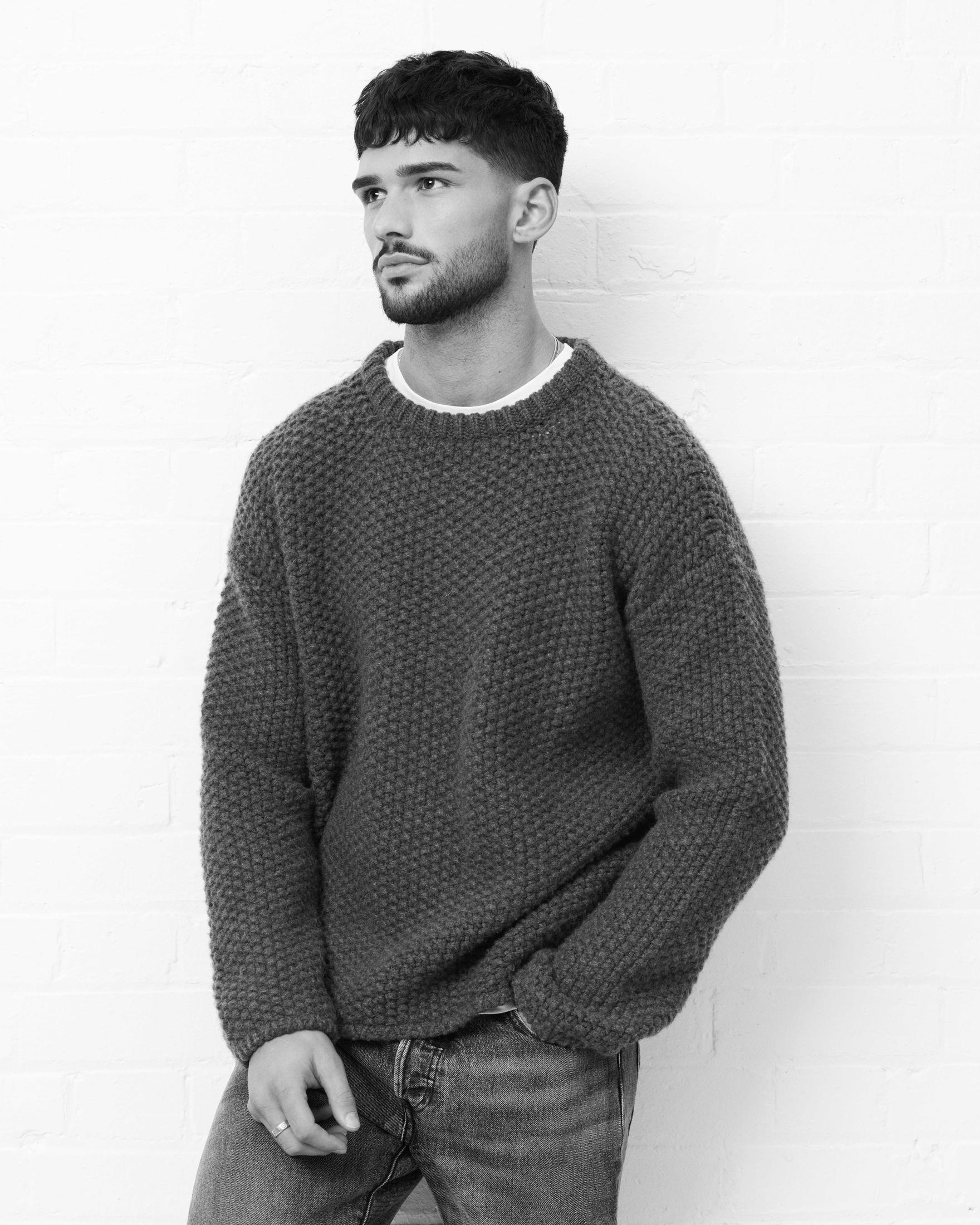 Mężczyzna w szarym swetrze i dżinsach, oparty o białą ceglaną ścianę.