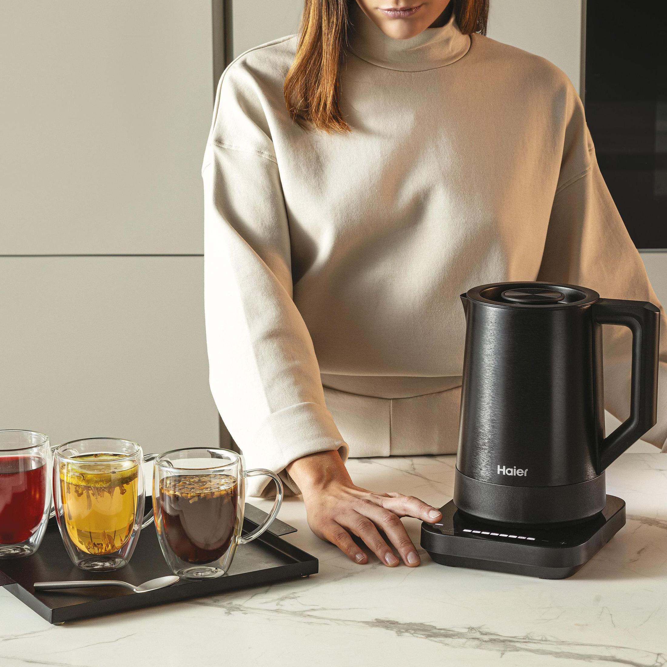 Kobieta stoi obok czarnego czajnika elektrycznego i trzech szklanek na marmurowym blacie.