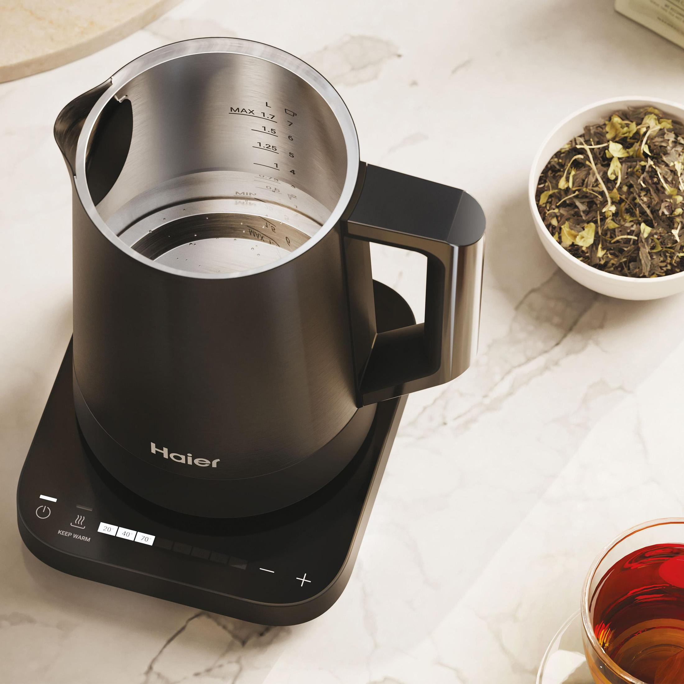 Czarny czajnik elektryczny na czarnej podstawie. Jest miska z ziołami i filiżanka herbaty.