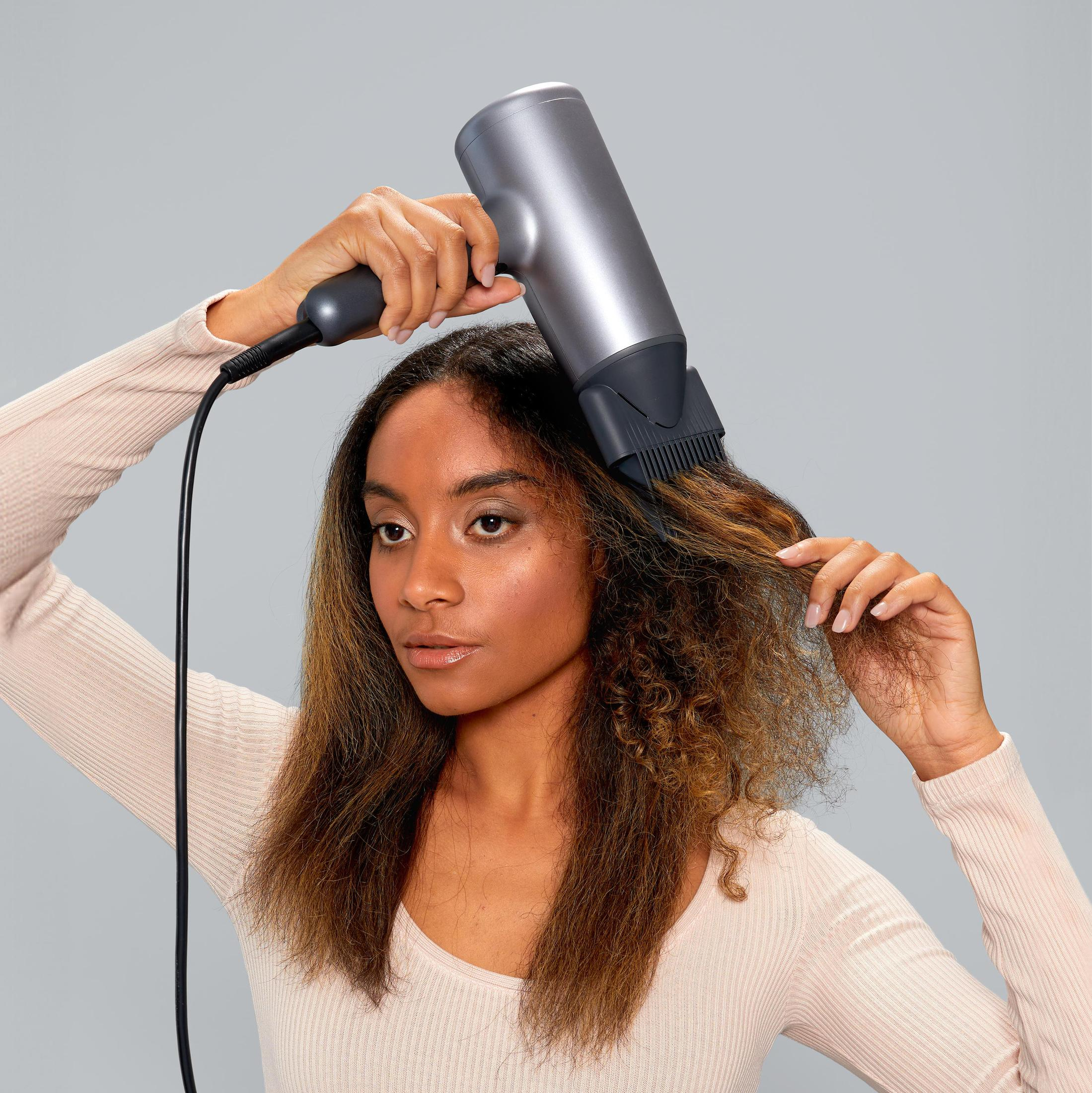 Kobieta używa szarej suszarki do włosów, susząc swoje kręcone włosy na jasnym tle.