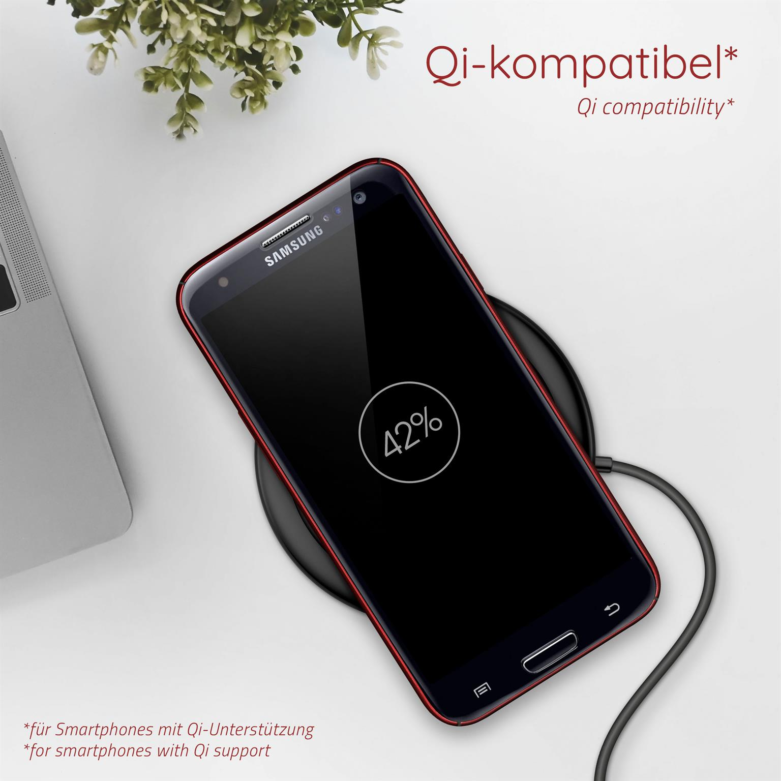 Smartphone auf einem schwarzen, kabellosen Ladegerät. Das Telefon zeigt 42%. Weiße Oberfläche mit Pflanze, Laptop.