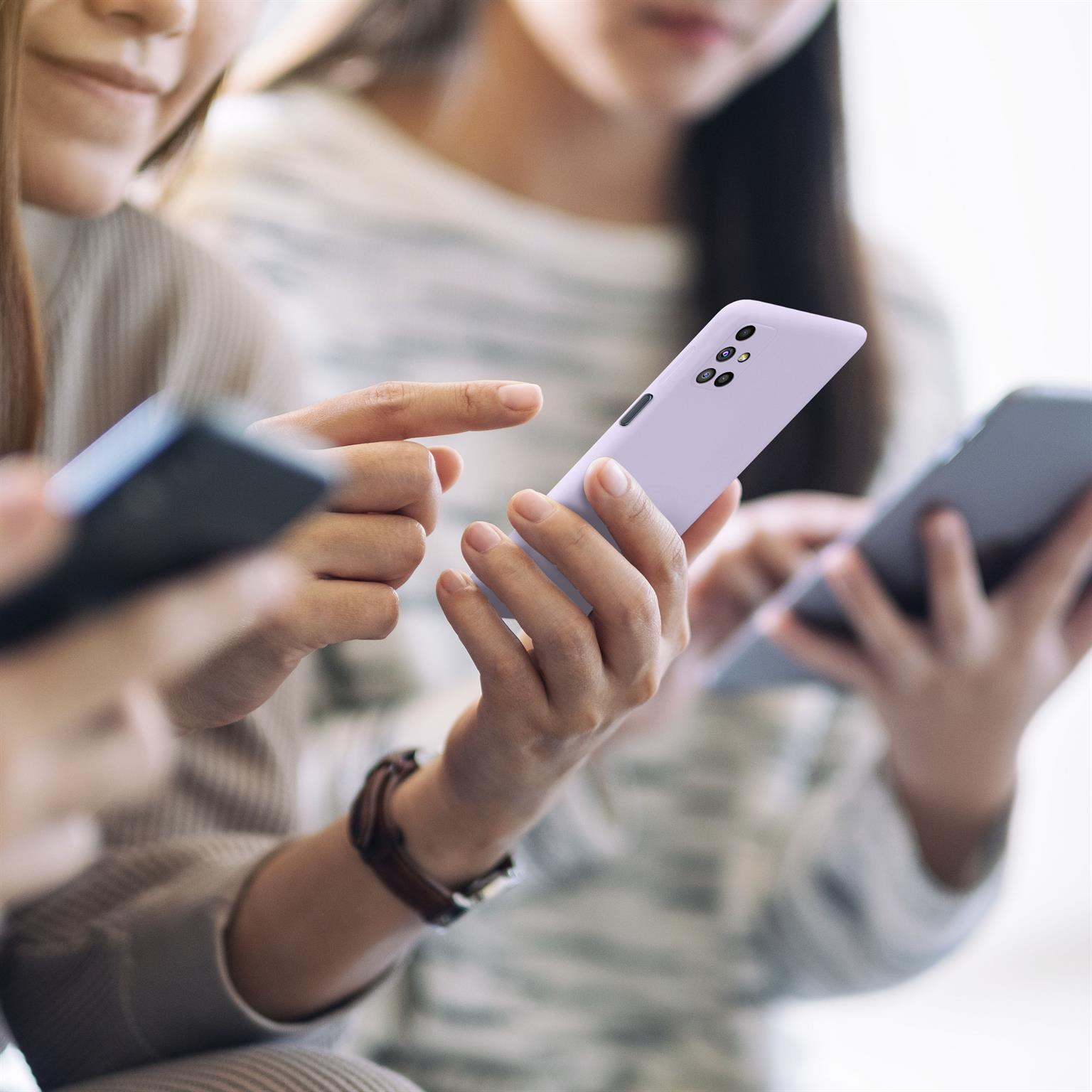 Drei Personen halten Telefone, eine mit lavendelfarbener Hülle. Alle schauen auf ihre Bildschirme. Verschwommener Hintergrund.