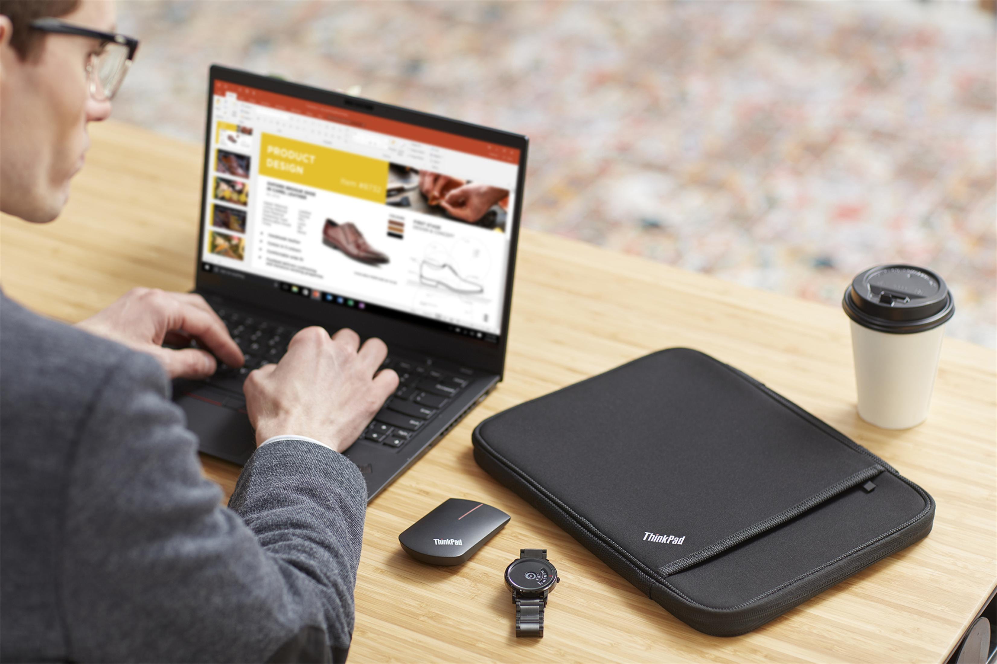 Uomo che lavora su un laptop, accanto a custodia ThinkPad, mouse, orologio e tazza di caffè. Scrivania in legno.