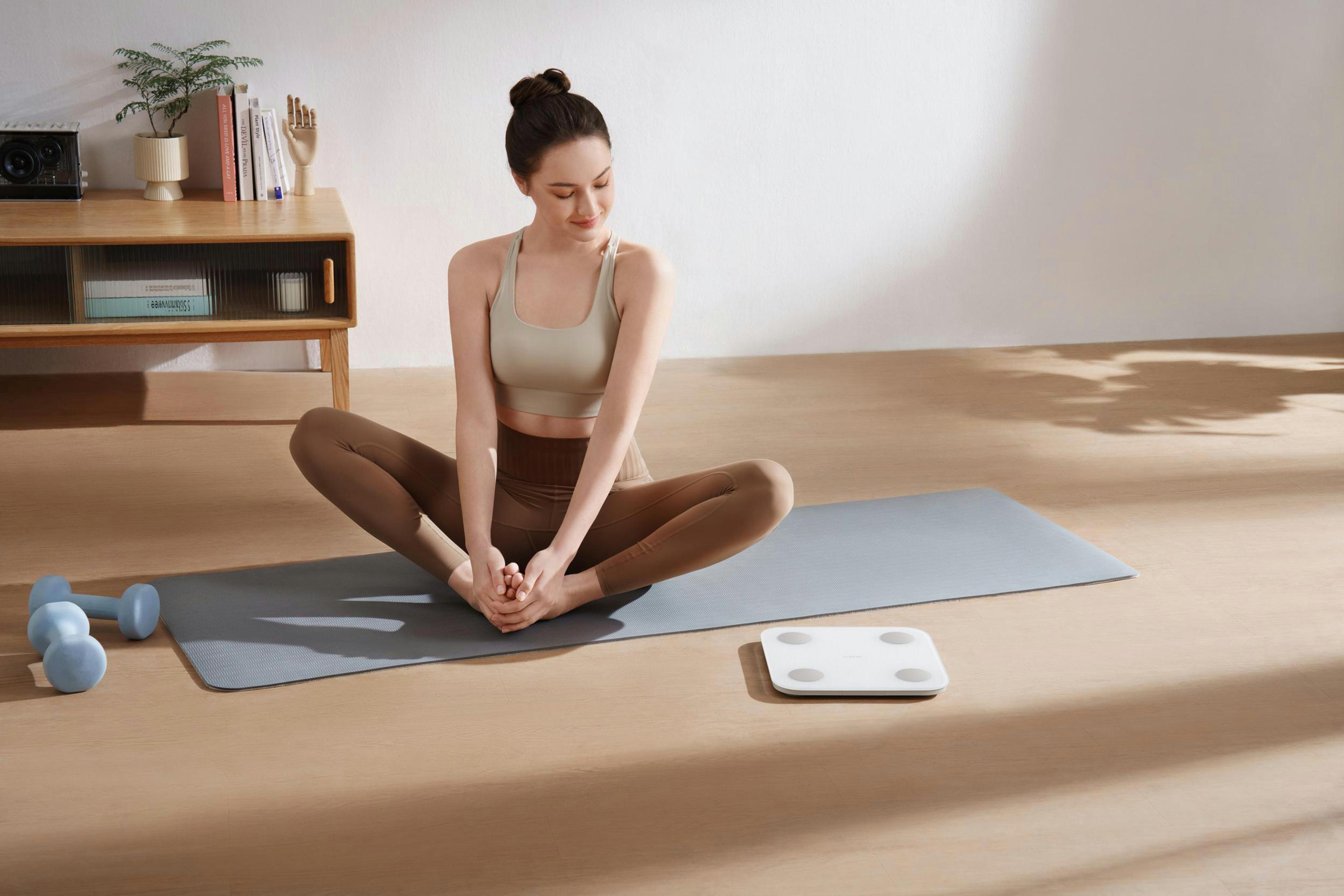Kobieta rozciąga się na macie do jogi. Na macie znajduje się biała waga. W pobliżu hantle i stół.