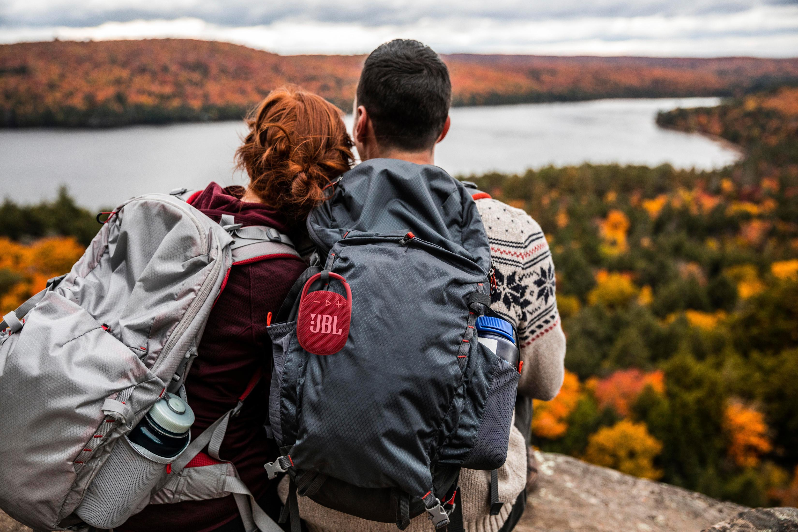 Pareja con mochilas mirando al lago, árboles otoñales al fondo. Altavoz JBL rojo en la mochila.