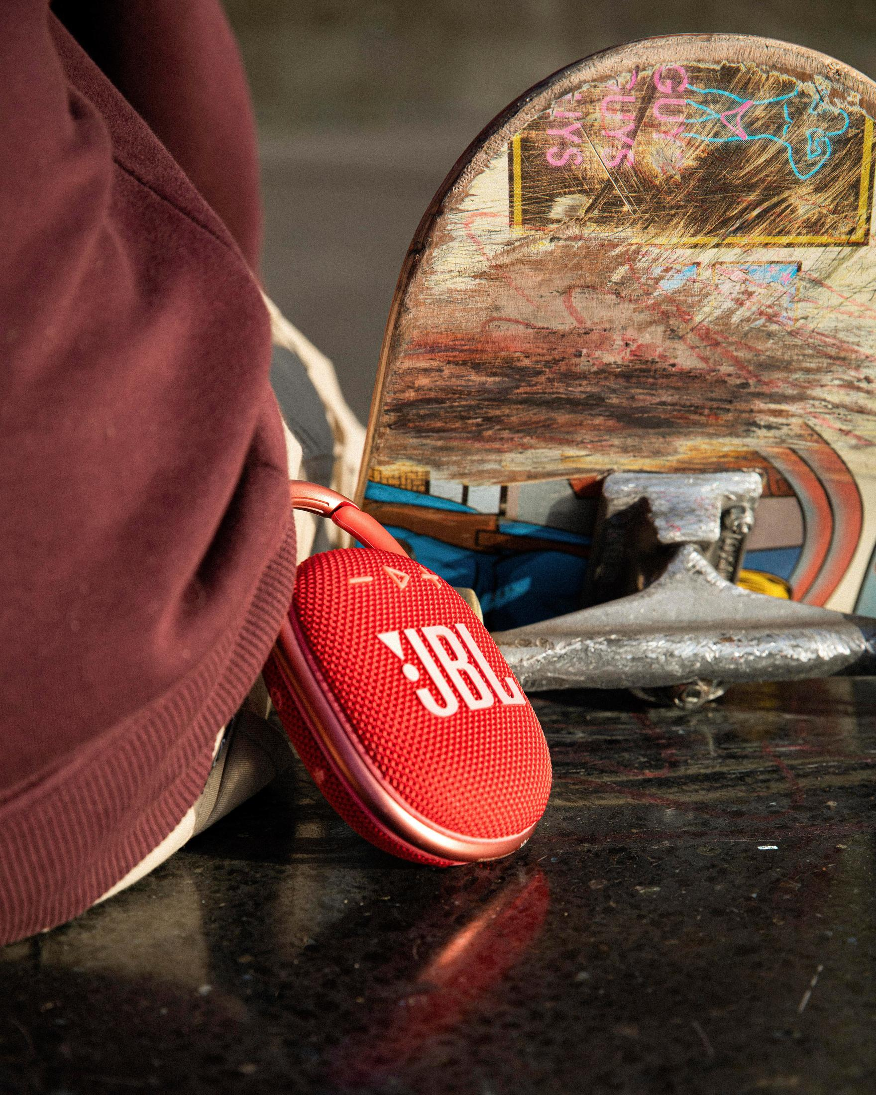 Altavoz JBL rojo junto a una patineta. La patineta se apoya en la persona.