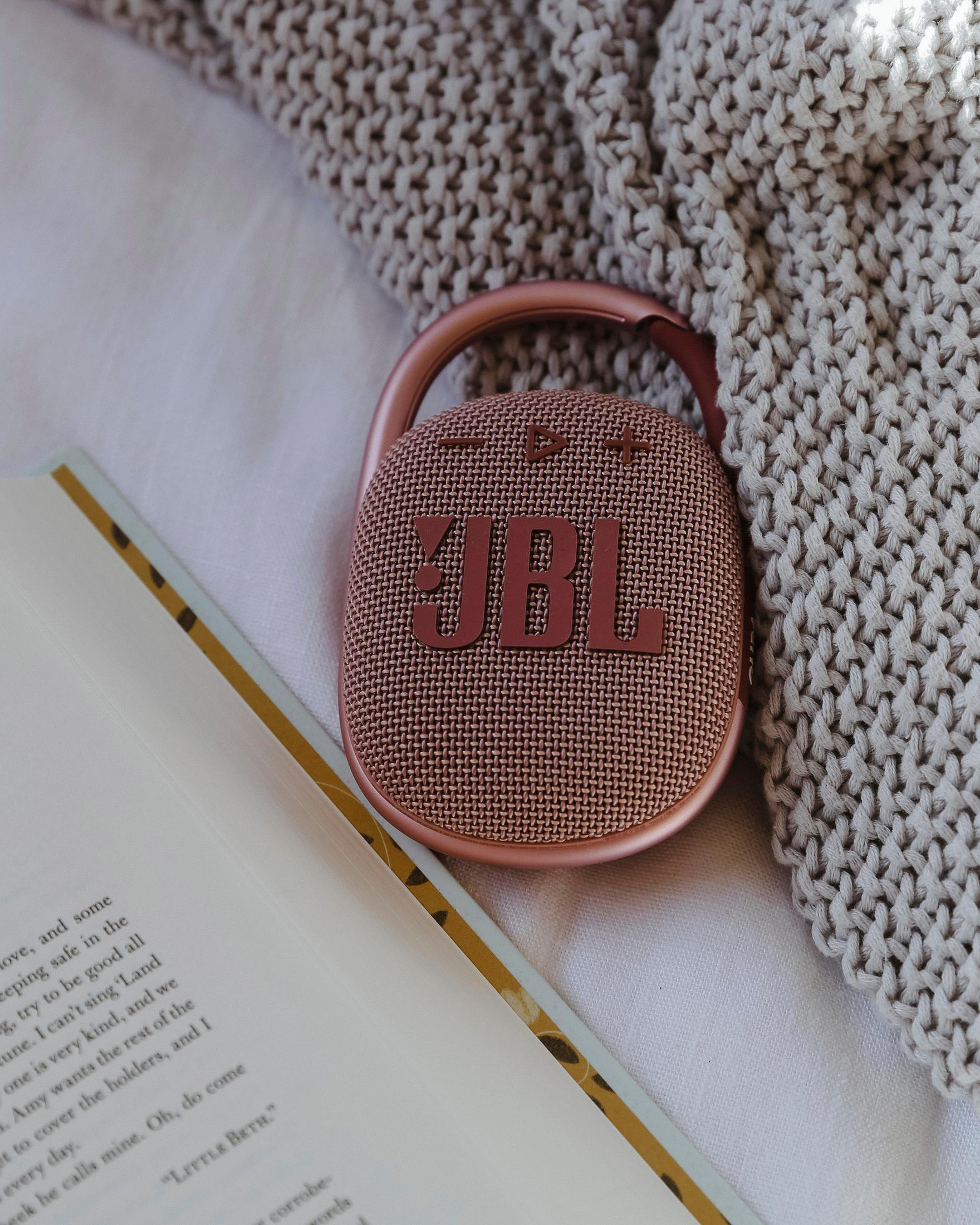 Głośnik JBL Clip, książka i koc na białej powierzchni. Głośnik jest różowy i ma logo JBL.