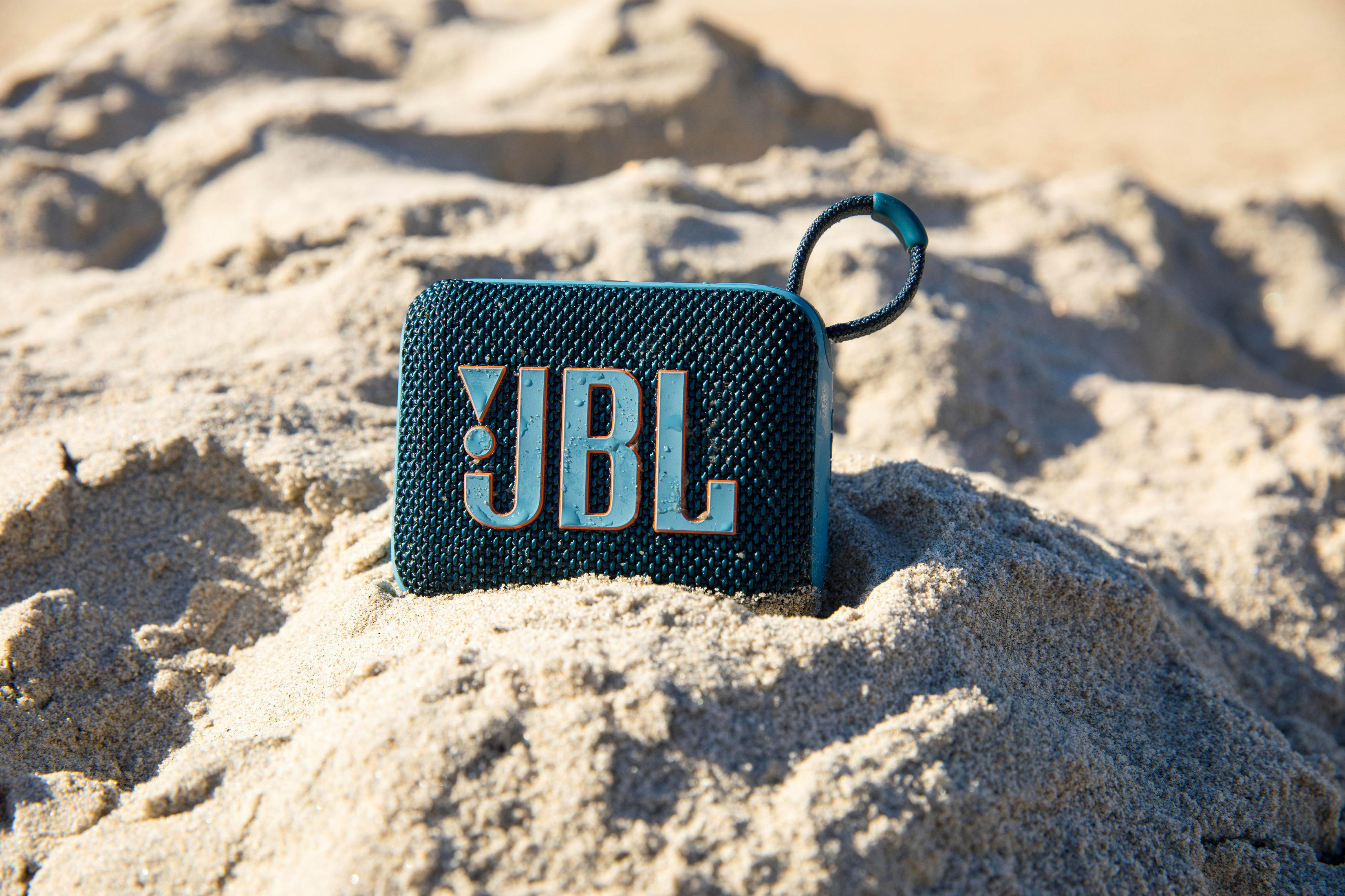 Altavoz portátil verde azulado en arena. Logotipo de JBL. Entorno de playa, día soleado.