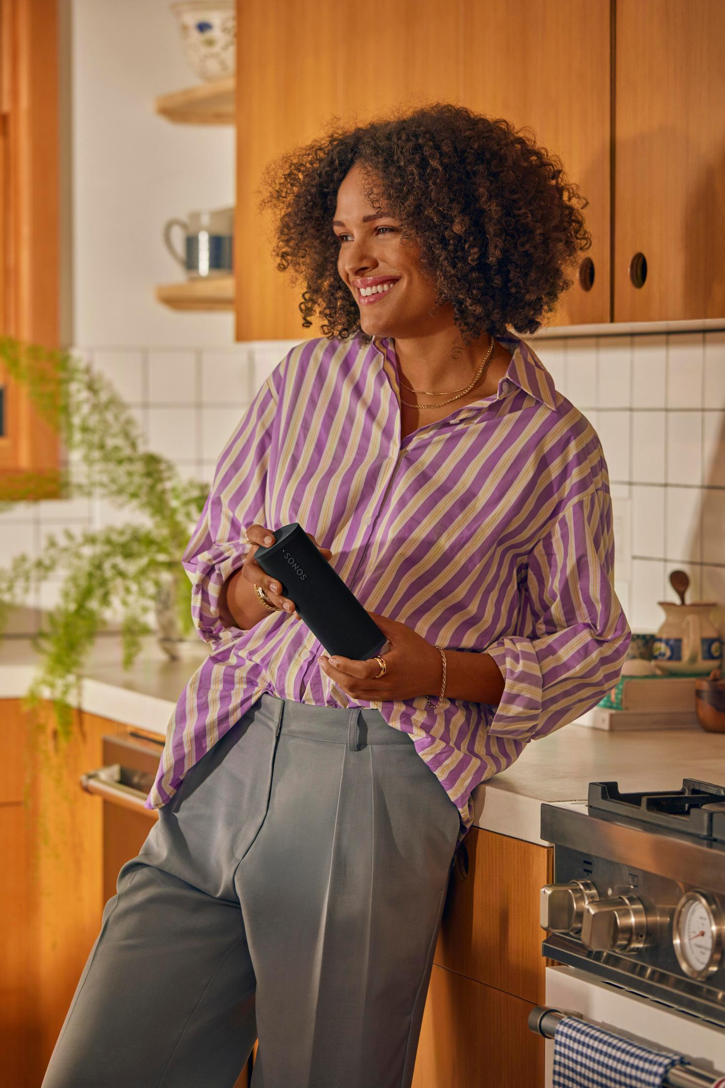 Donna con camicia a righe che tiene in mano un altoparlante nero in cucina. Sorride, appoggiata al bancone.