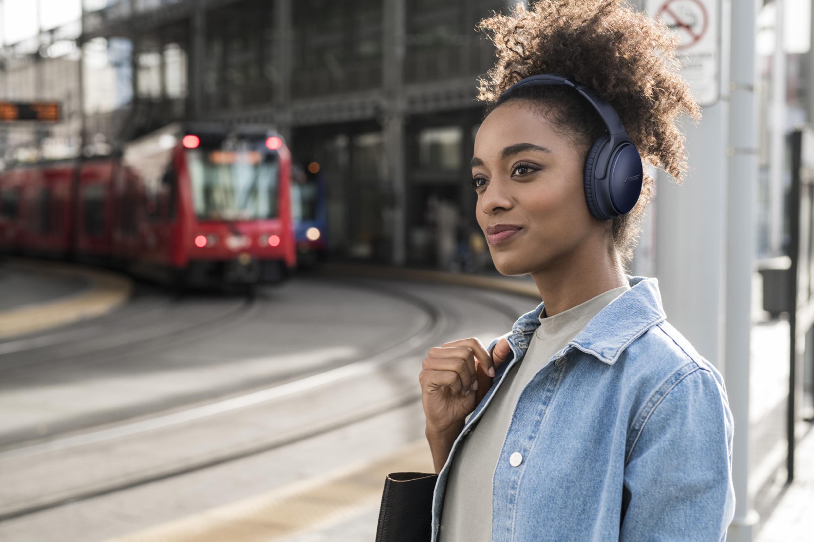 Frau in Jeansjacke mit Bose-Kopfhörern, am Bahnhof stehend.