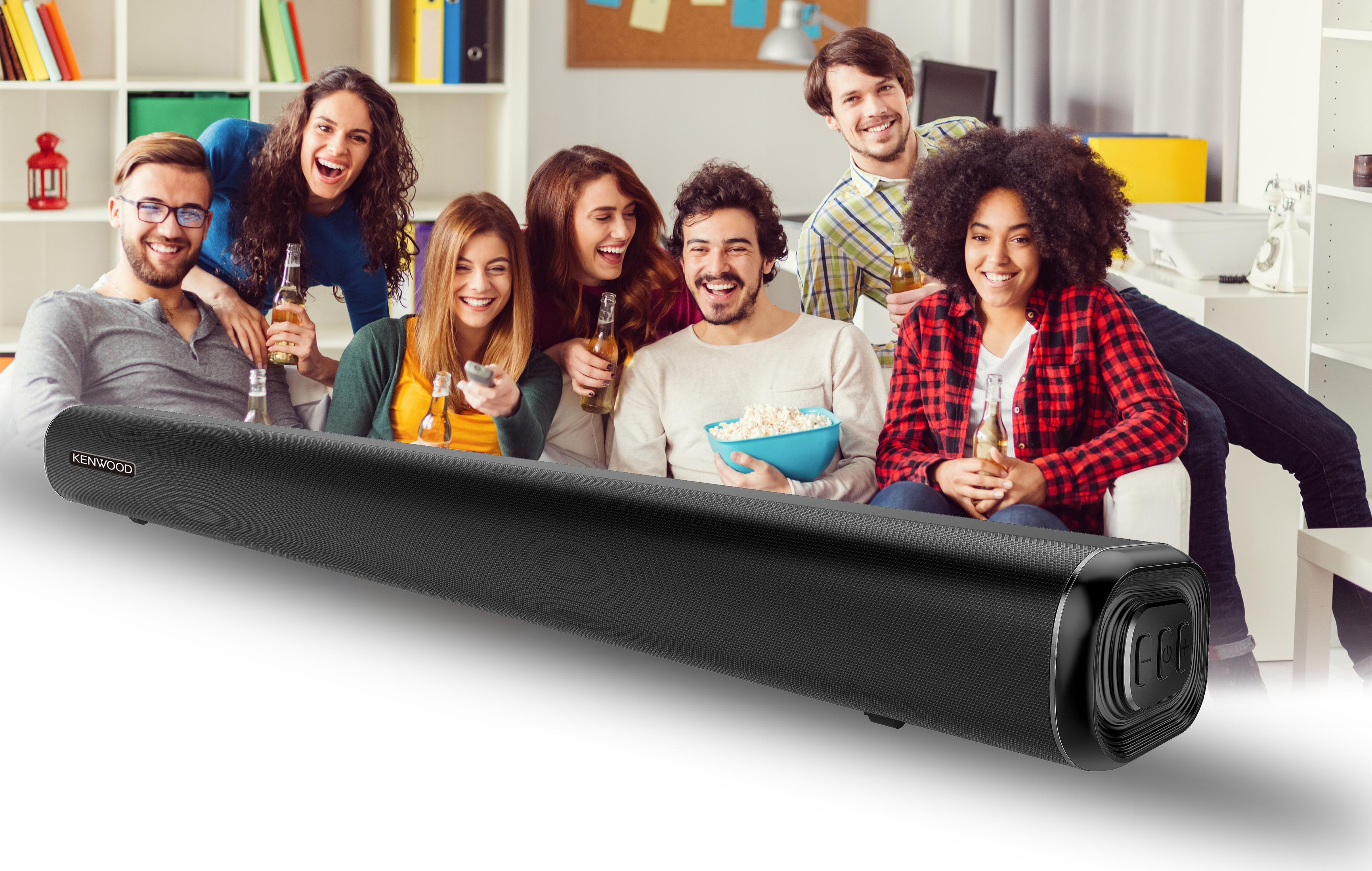 Un gruppo di persone sedute su un divano sorridono, tenendo in mano bevande e snack, vicino a una soundbar Kenwood.