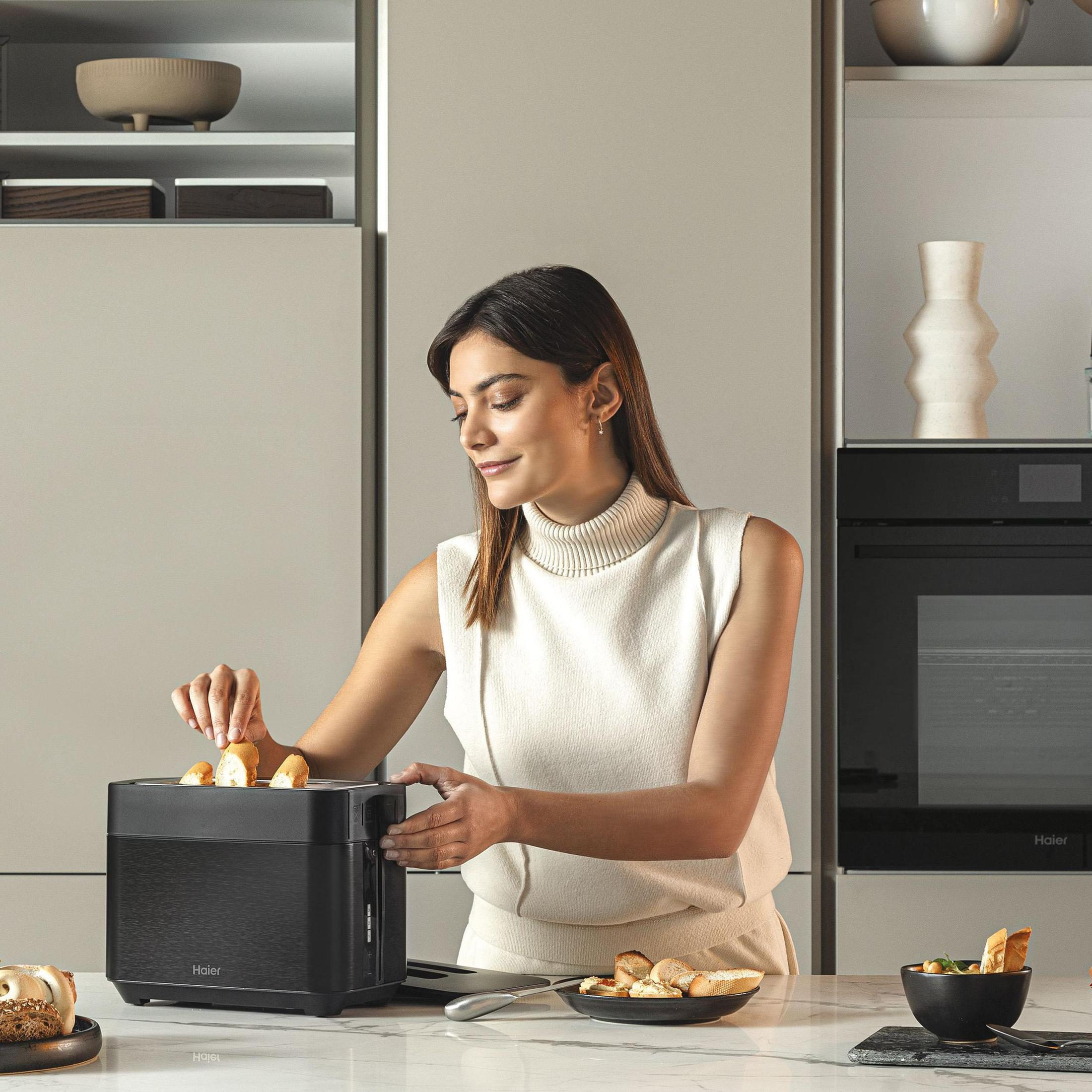 Kobieta wkłada chleb do czarnego tostera. Otoczenie kuchni z talerzami i akcesoriami.