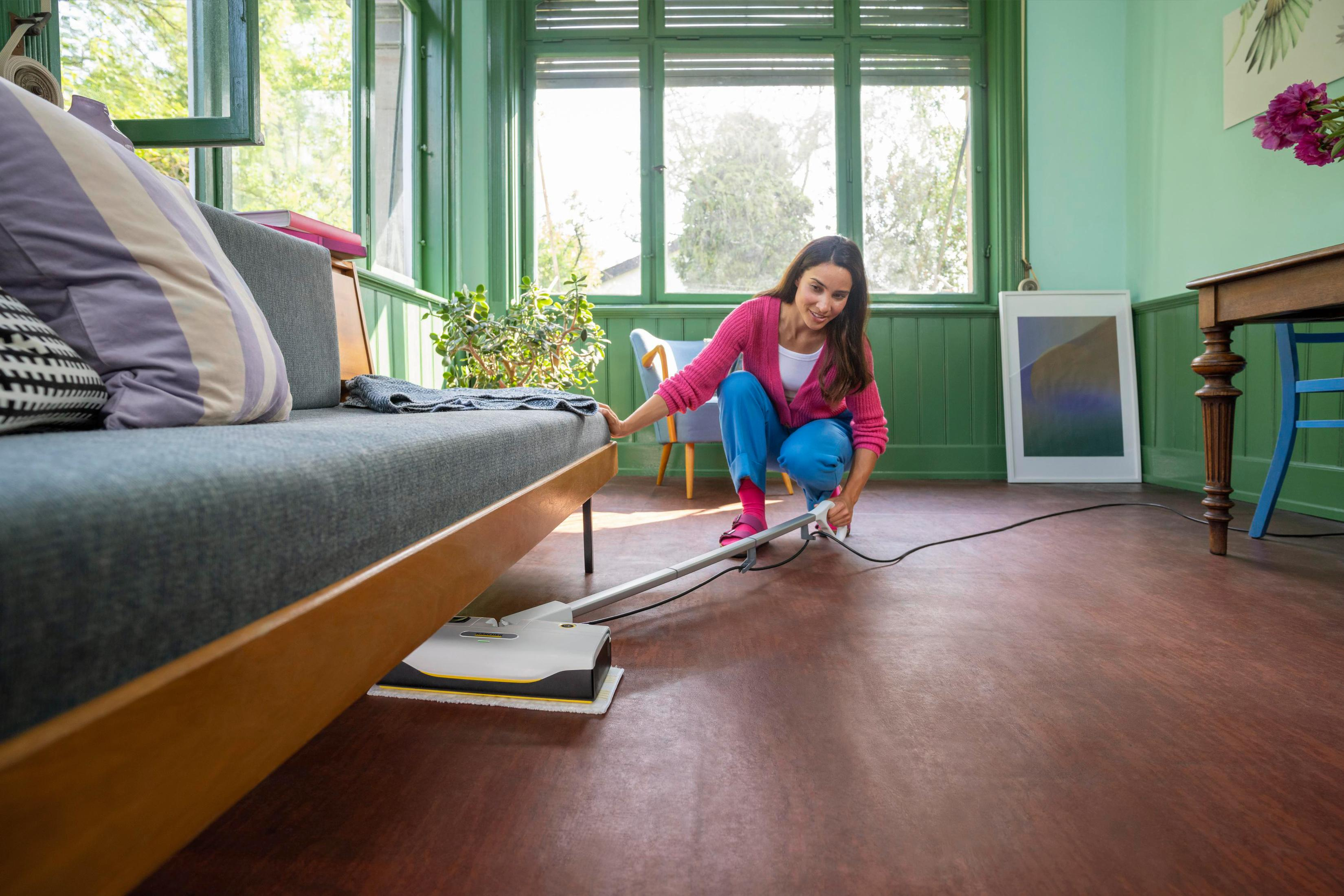 Kobieta czyści drewnianą podłogę za pomocą myjki do podłóg. Pokój ma zielone ściany i sofę.