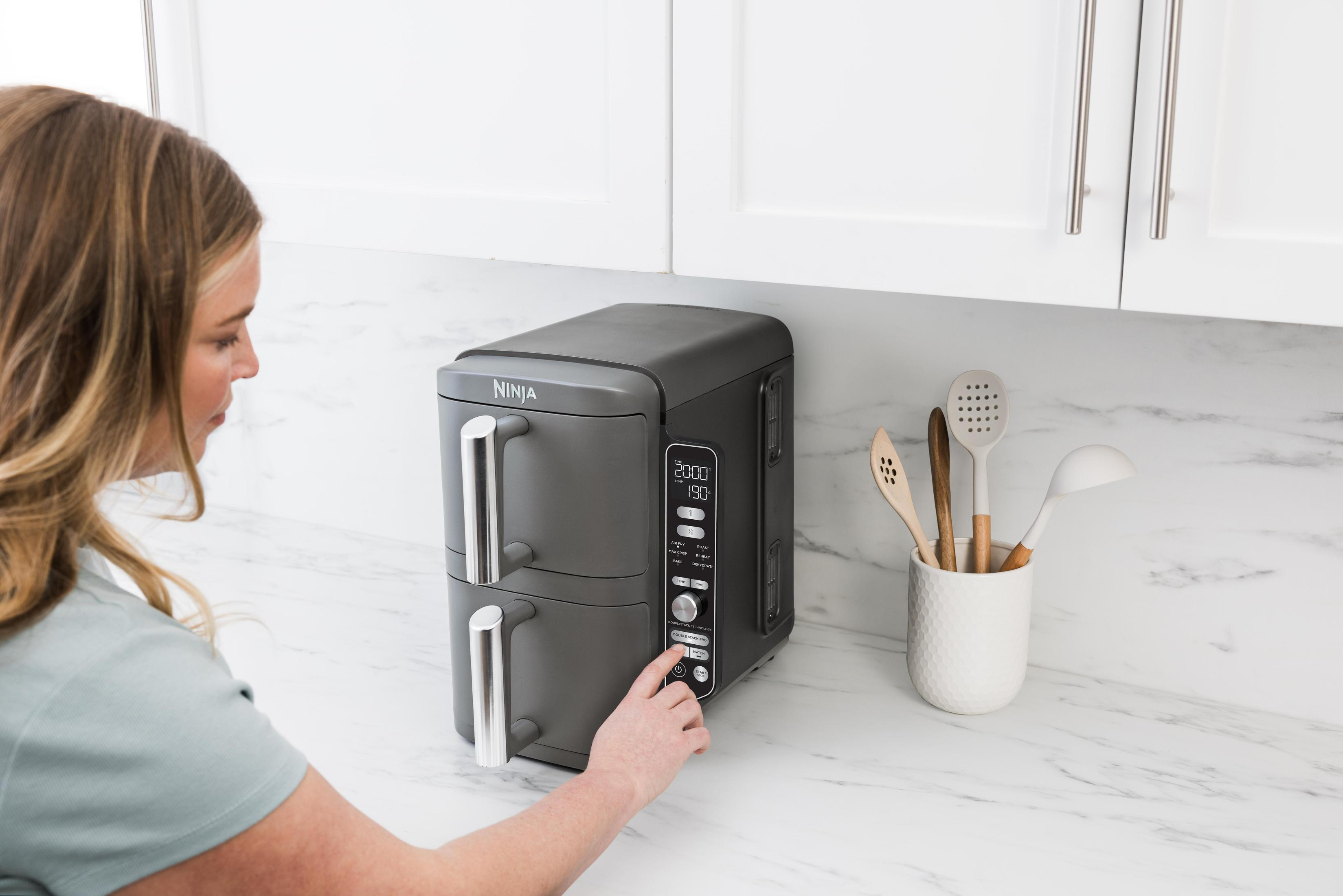Kobieta naciska przycisk na szarej frytkownicy powietrznej Ninja, na białym blacie, obok białej szafki i przyborów kuchennych.