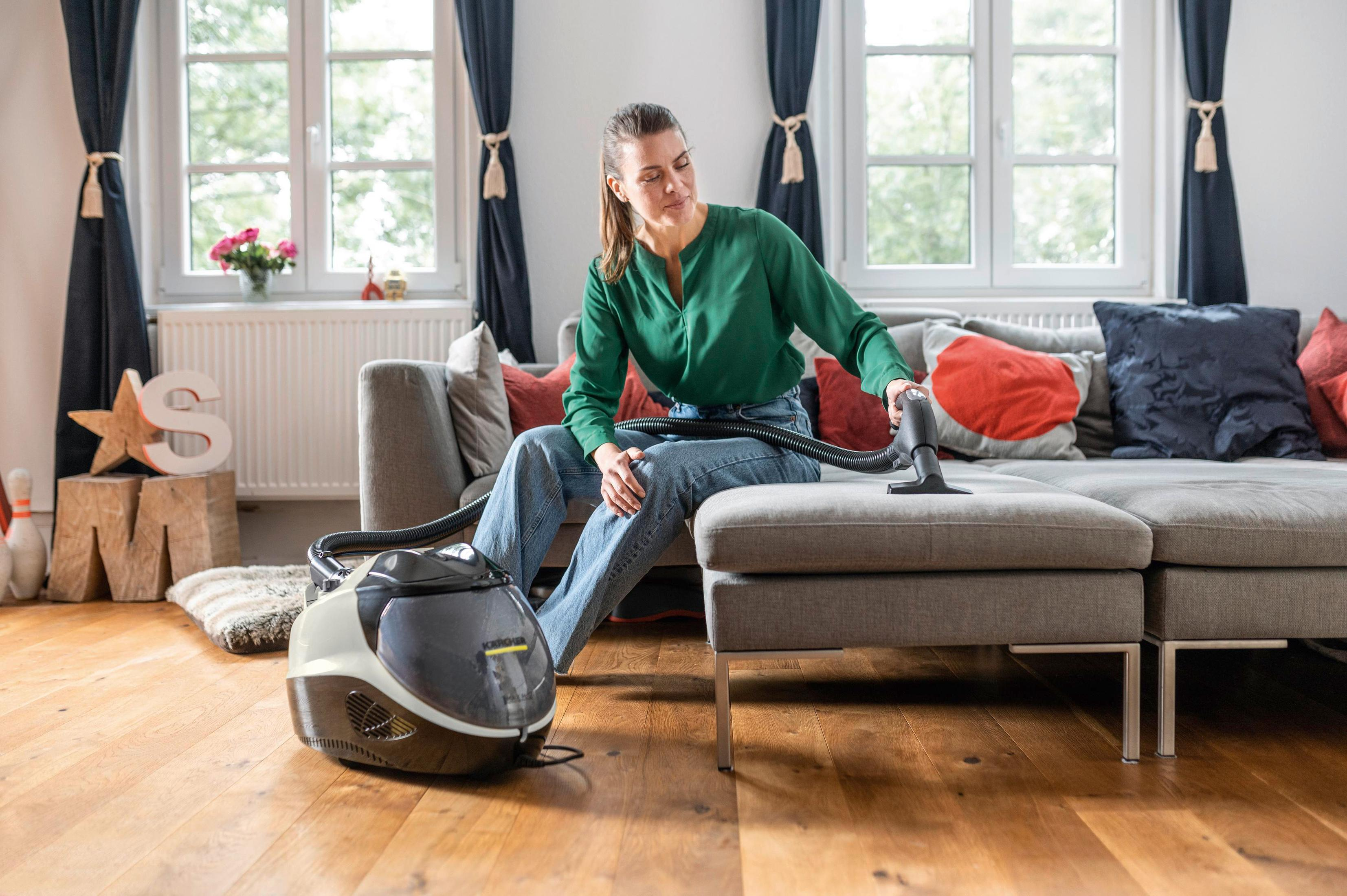 Kobieta sprzątająca szarą kanapę odkurzaczem w salonie z drewnianymi podłogami i oknami.