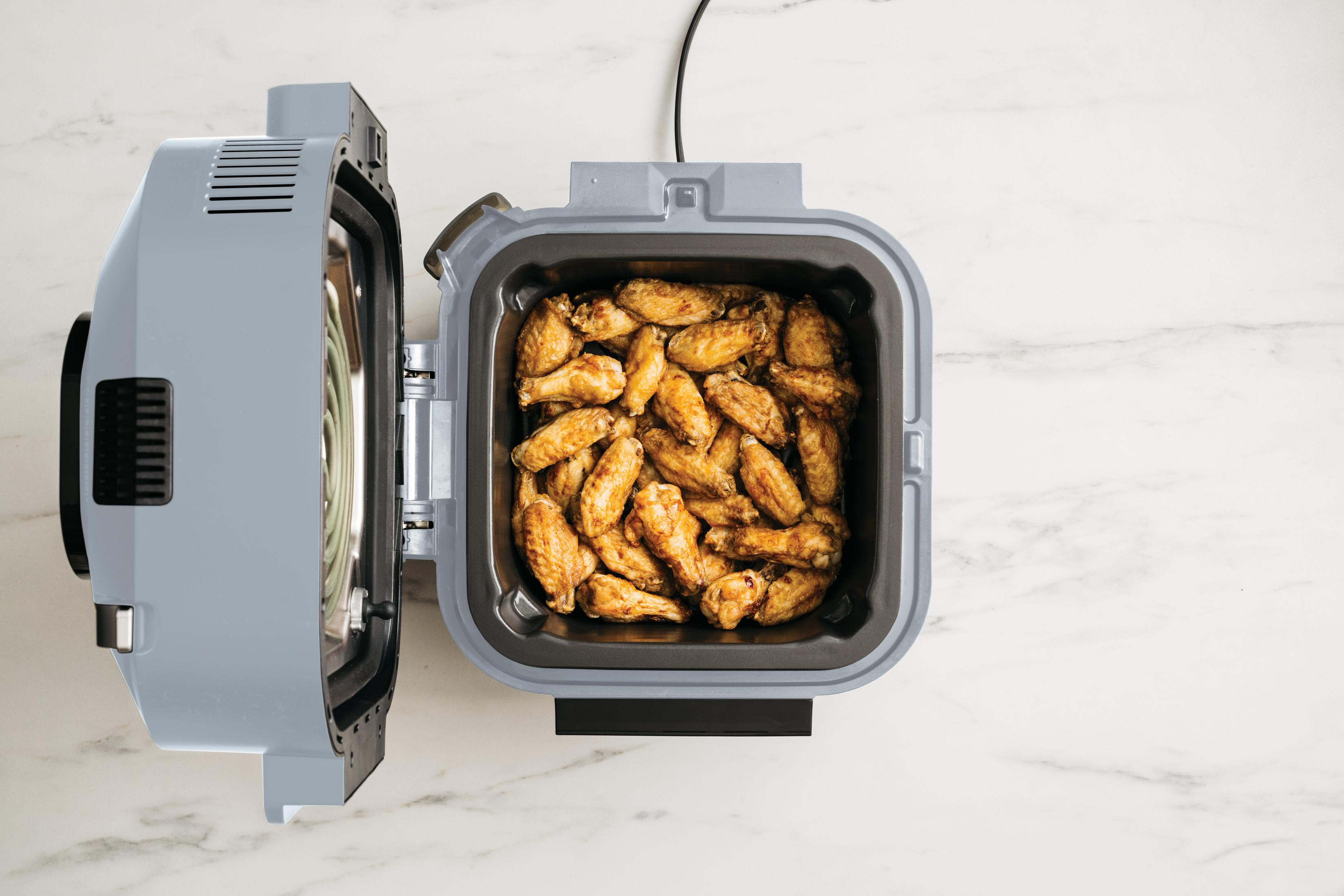 Air fryer open, full of cooked chicken wings, on white marble.