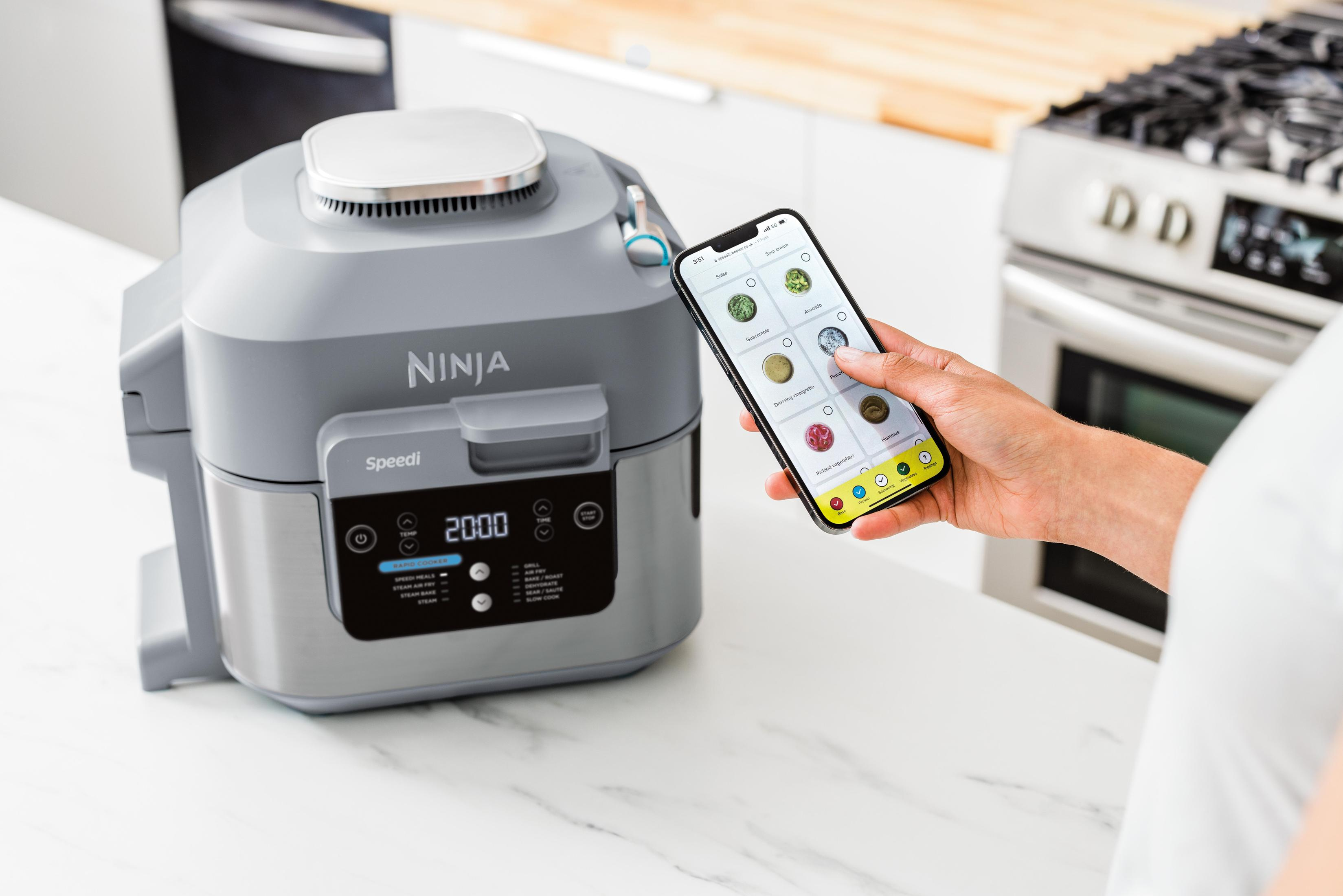 A Ninja Speedi cooker with a phone showing an app interface. The screen shows food options.
